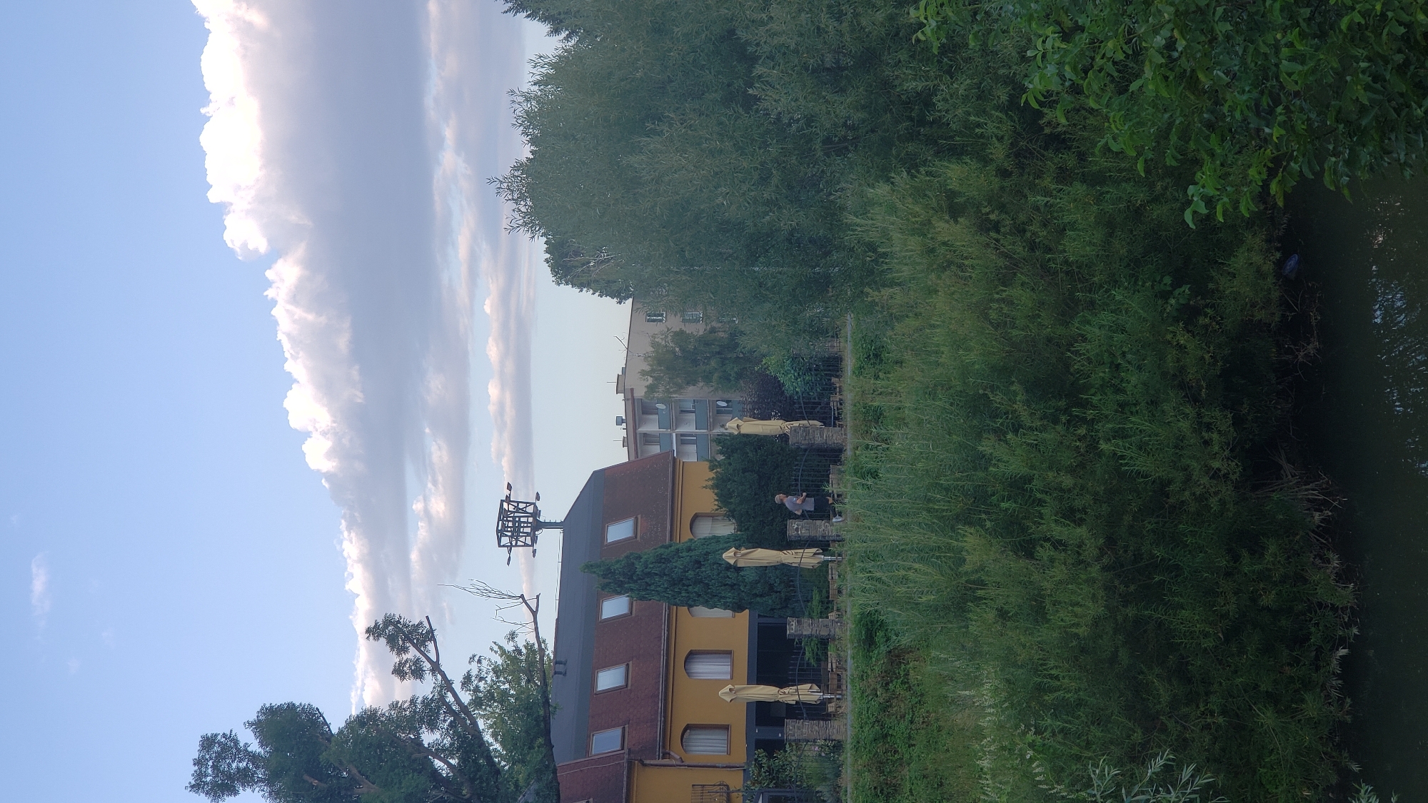 The image shows a view of a building with a yellow facade and a dark roof with several dormer windows. In front of the building, there are several closed umbrellas, suggesting an outdoor seating area. A metal fence separates this area from a grassy embankment. A person is jogging along the embankment, wearing a grey t-shirt and dark shorts. Behind the yellow building, there are other buildings, including a residential building with a light-colored facade and balconies. To the right of the yellow building, a large, dense green tree dominates the scene. In the foreground, there is a body of water, likely a river or canal, with lush green vegetation growing along its banks. The sky is blue with scattered white clouds. The scene appears to be in a suburban or park-like area within the city of Prešov, Slovakia, possibly near a restaurant or hotel with an outdoor terrace.