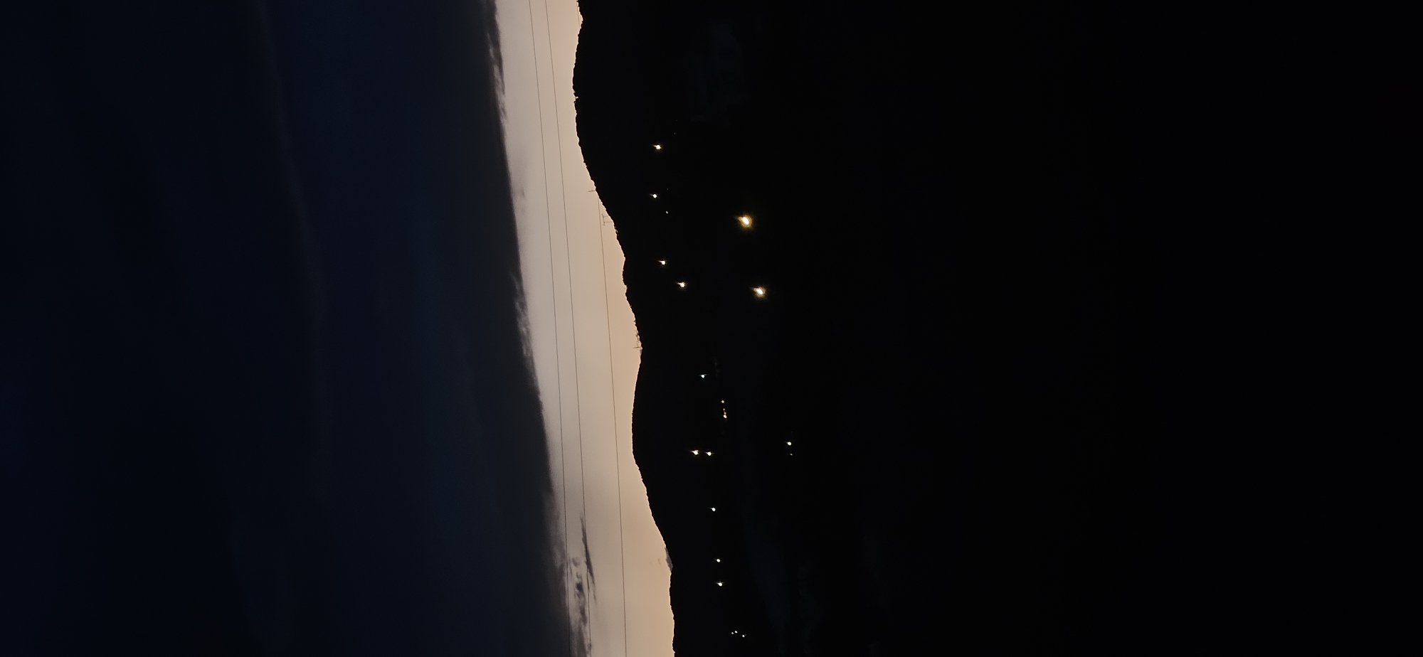 The image, viewed in its correct orientation, captures a nocturnal or late twilight landscape featuring a silhouetted mountain range against a partially lit sky. The sky transitions from dark at the top to a lighter, pale orange or grey hue near the horizon, indicative of dusk or early night. A prominent, dark band of clouds stretches across the middle portion of the sky, above the lighter horizon line.
Below the cloud band, the distinct silhouette of mountains or hills forms a jagged, undulating line. Along the visible slope of these mountains, a winding trail of small, bright, warm-toned lights is visible, suggesting a road or path ascending the terrain. These lights glow softly against the deep black of the mountain and the surrounding darkness. Several faint, parallel lines, likely power lines, are discernible stretching horizontally across the sky, running above the mountain silhouette.
The scene conveys a sense of vastness and tranquility, characteristic of a mountainous area at night. There are no visible people or specific events taking place, only the static landscape. No text is visible within the image. The setting aligns with the mountainous terrain found in locations like Momin Prohod, Bulgaria.