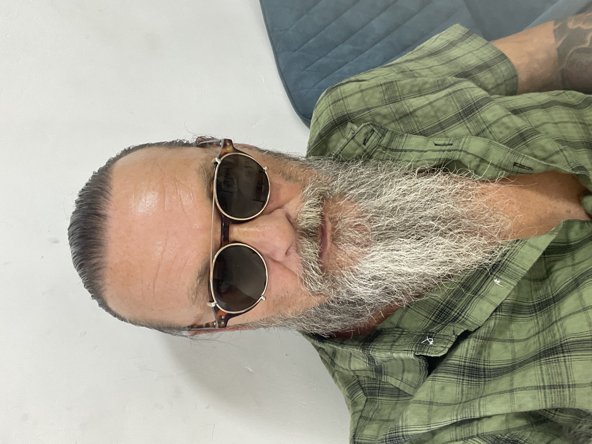 The image is a selfie of a man with a long, gray beard, wearing sunglasses with round, tortoise-shell frames. He has his dark hair pulled back and is wearing a green plaid short-sleeved shirt. He appears to be indoors, in front of a white wall, with a blue upholstered surface to his right, possibly a sofa. His left arm is visible, showing a tattoo. The scene does not contain information to determine a specific location within Netanya, Israel.