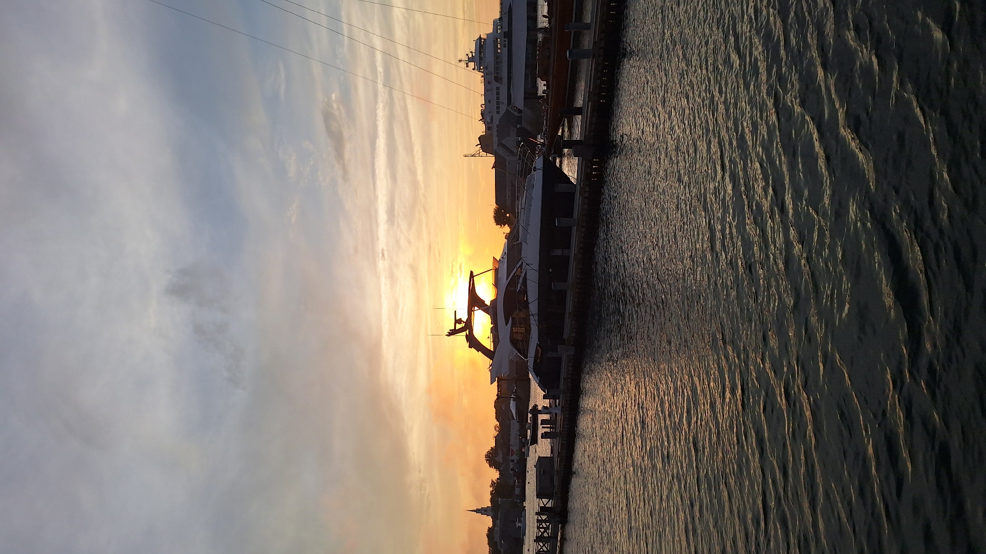 The image depicts a harbor scene in Nantucket, Massachusetts, during sunset. Several yachts are docked along a wooden pier in the foreground, their sleek lines silhouetted against the warm orange and yellow hues of the setting sun. Beyond the yachts, the Nantucket town skyline is visible, characterized by a white steeple of a church. The water in the foreground is rippled, reflecting the colors of the sky. In the background, a large ferry is docked. The sky is filled with scattered clouds, some of which are tinged with orange from the setting sun. The overall atmosphere is peaceful and serene.