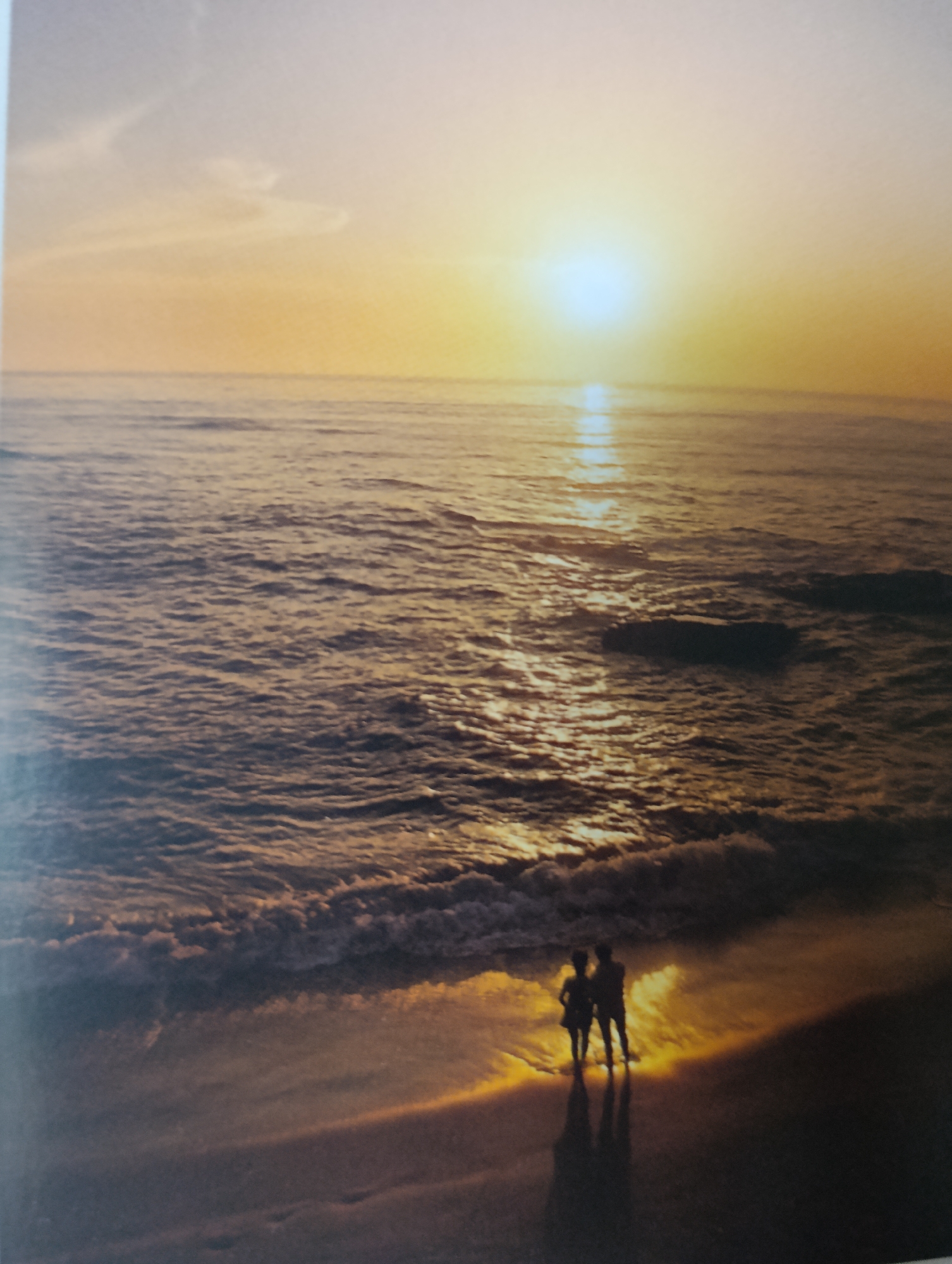 An aerial, full shot captures a tranquil sunset over a calm ocean with two figures standing on the wet sand at the shoreline. The sun is a bright orb of light on the horizon, casting a golden sheen on the water's surface. The water is a deep blue-grey, with gentle waves lapping the shore. The two figures, silhouetted against the setting sun, stand close together, their reflections shimmering on the sand. The beach itself is dark and sandy, with the golden light from the sun creating a stark contrast. The scene evokes a sense of peace and romance, with the natural beauty of the sunset and the intimacy of the couple.