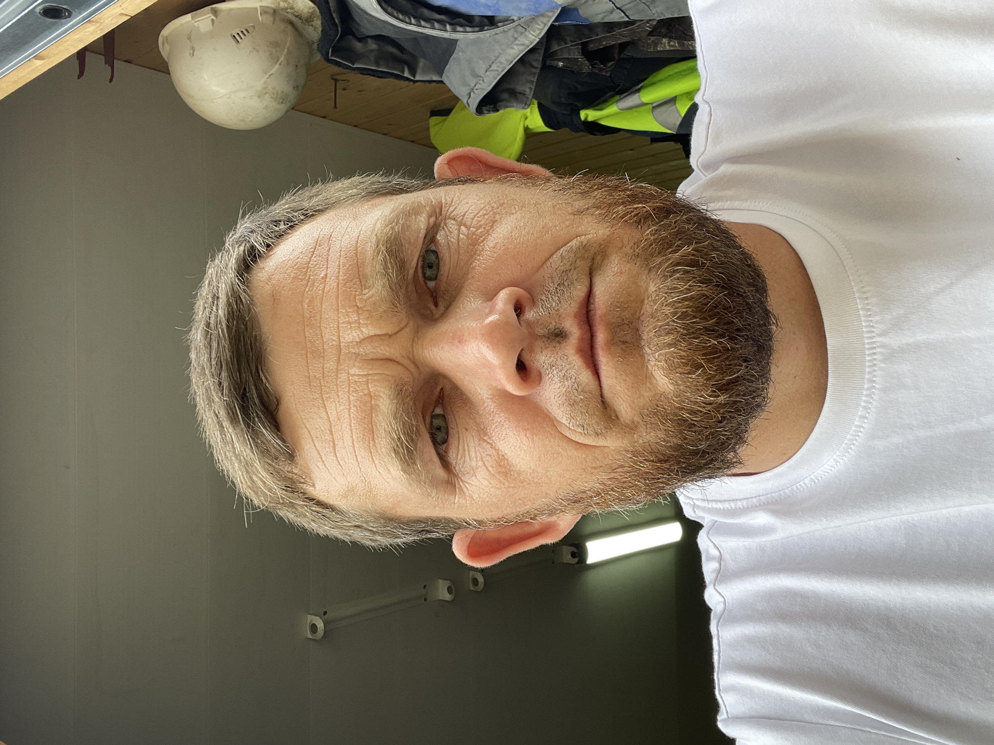 A close-up, head-and-shoulders selfie of a light-skinned man with short, sandy brown hair, a trimmed beard of the same color, and light green eyes. He is wearing a white t-shirt. The background consists of a grey ceiling with fluorescent lights and some wooden paneling on the right side. A white hard hat is visible hanging on the wall, along with some work jackets, one of which is a high-visibility lime green. The image appears to be taken indoors, possibly in a workshop or garage. There is no indication of the specific location in Krasnodar, Russia.