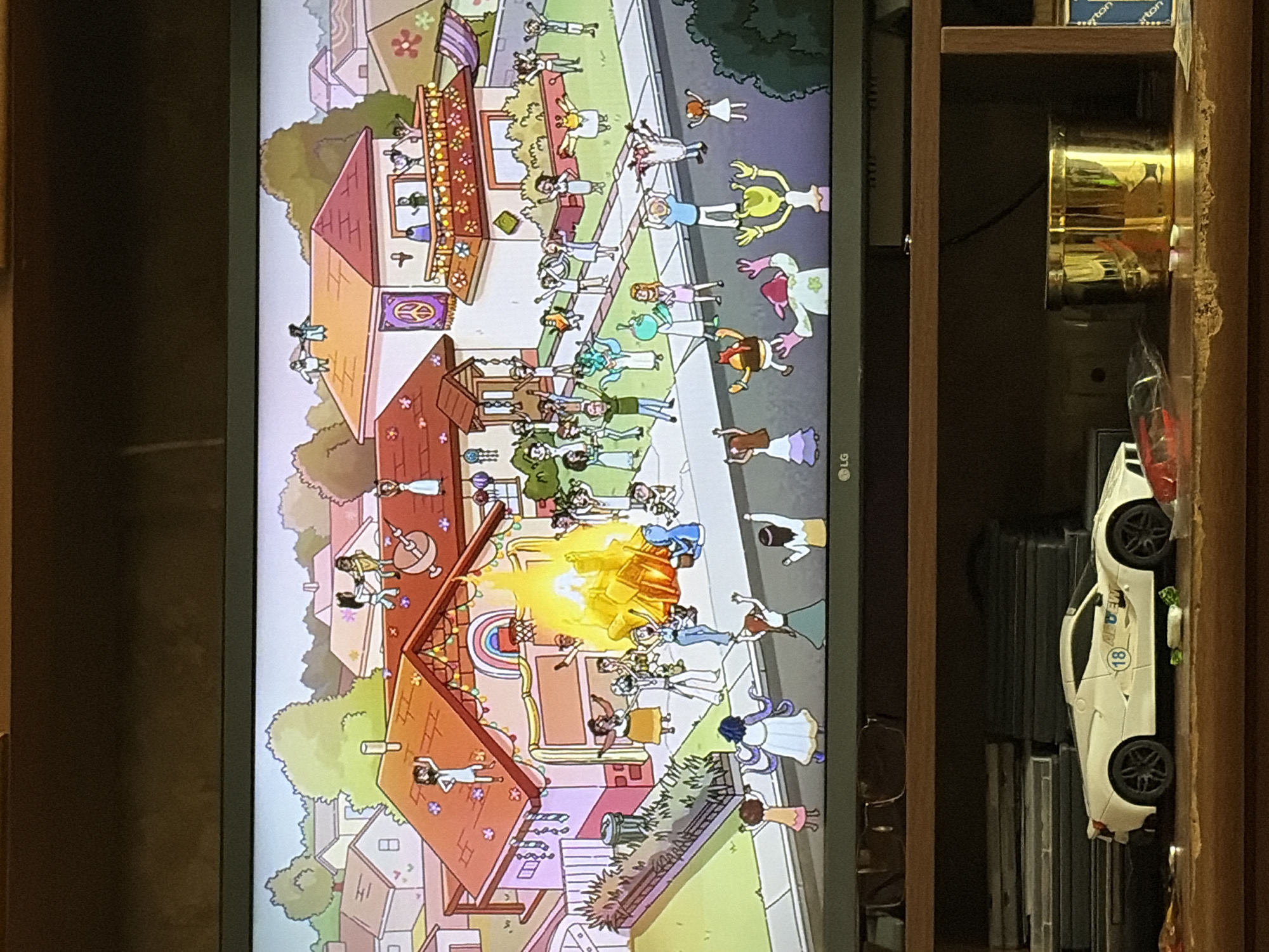 The image depicts a cartoon scene of a suburban neighborhood with many people gathered on rooftops and in the street. A large bonfire is burning in front of a house, and many of the individuals are raising their arms in celebration. The houses are colorful and have various decorations, including flower motifs and peace signs. In the foreground of the image, on a wooden shelf below the television screen, sits a white toy sports car and a gold cylindrical container. The toy car has the number 18 on a blue circular sticker and some decals on its side. The gold container is highly reflective.