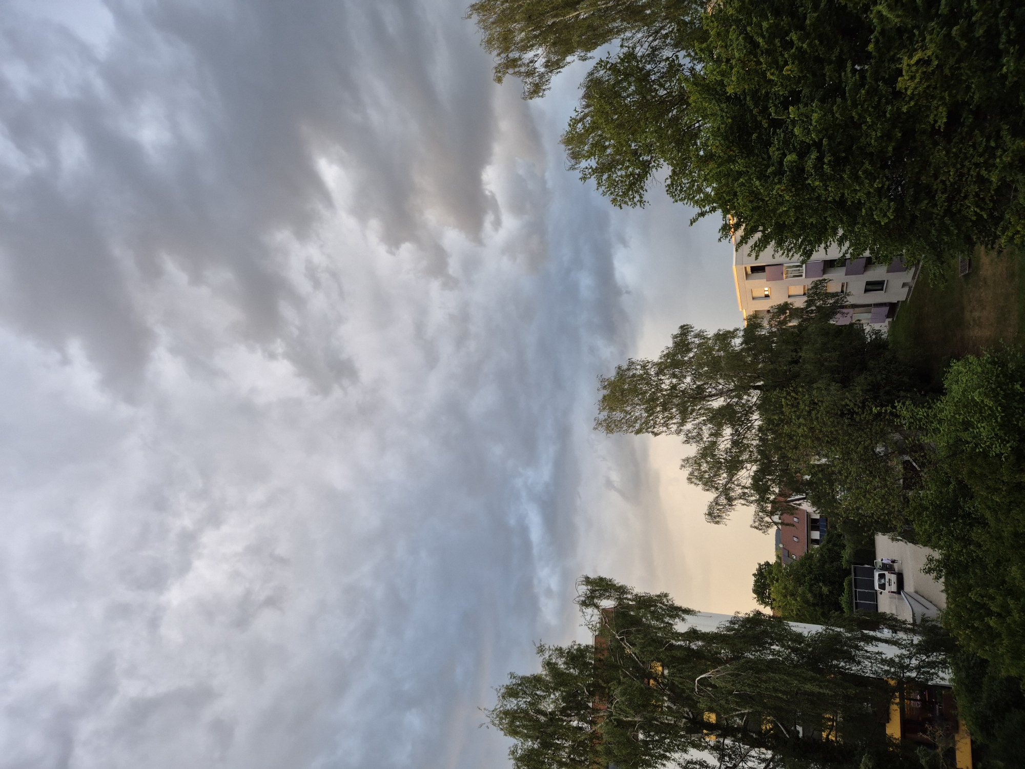 The image displays an outdoor residential scene under a heavily overcast sky. The sky, occupying the upper two-thirds of the frame, is filled with a dense cover of grey and white stratocumulus clouds, exhibiting subtle variations in illumination with brighter patches and darker grey sections. Towards the lower horizon, particularly on the left and center, a soft, warm, peachy-orange glow is visible beneath the clouds, indicating a low sun obscured by cloud cover.
The foreground and mid-ground are dominated by lush green foliage from numerous trees and bushes. On the far left, a tall deciduous tree with light green, somewhat drooping leaves partially obstructs a multi-story building featuring a yellow lower section and white upper facades. In the mid-ground, a cluster of darker green deciduous trees forms a dense canopy. On the right, parts of another tall tree with light green foliage are visible, framing the scene.
Between the trees and foliage, several buildings are discernible. In the central background, beyond a paved parking area, a house with a reddish-brown tiled roof is visible, with what appear to be dark rectangular panels (possibly solar) mounted on the roof. In the mid-right, a multi-story residential building with white walls and distinctive light purple balconies is prominent. One window on this building shows an interior light source.
In the paved parking area, a white van or utility vehicle is parked, facing away from the viewer. A small person, possibly a child or short adult, wearing dark clothing, is standing near the rear of the white van. Green grassy areas are visible between the buildings and planted foliage. The overall lighting is diffused and soft, characteristic of an overcast late afternoon or early evening in a residential area within an unknown city or town.