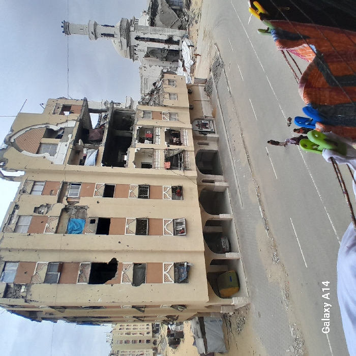 The image captures a street scene in Khan Yunis, Palestinian Territory, under an overcast sky, viewed from an elevated position with laundry partially obscuring the lower right foreground. The central subject is a heavily damaged multi-story residential building with a beige facade, exhibiting significant structural compromise including large sections of missing exterior walls revealing internal rooms and rebar, particularly on its upper floors and right side. Numerous smaller impact marks are visible across the building's surface. Ground-level arches provide shelter for two parked vehicles, one yellow-green and one light-colored. To the right and behind this building stands a partially destroyed mosque, characterized by a visible white minaret and a damaged dome structure, surrounded by rubble. An asphalt road with a visible centerline runs horizontally across the midground. Four individuals are present on the road: one adult male standing, and another adult with two small children standing near them. In the left background, another multi-story building appears damaged, and piles of debris and rubble are scattered across the landscape.