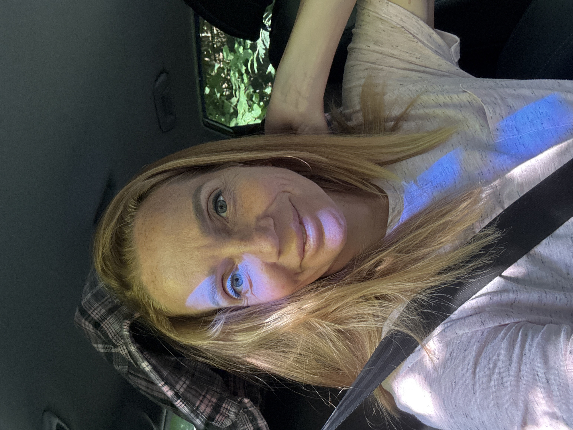 A woman with blonde hair and blue eyes is sitting in the passenger seat of a car. She is wearing a light pink t-shirt and a seatbelt. The sun is shining through the window, casting shadows and light patterns on her face and clothing. Outside the car window, green foliage is visible, indicating a natural, possibly wooded, environment. The interior of the car appears dark. The image is a selfie, taken from a front-facing perspective.