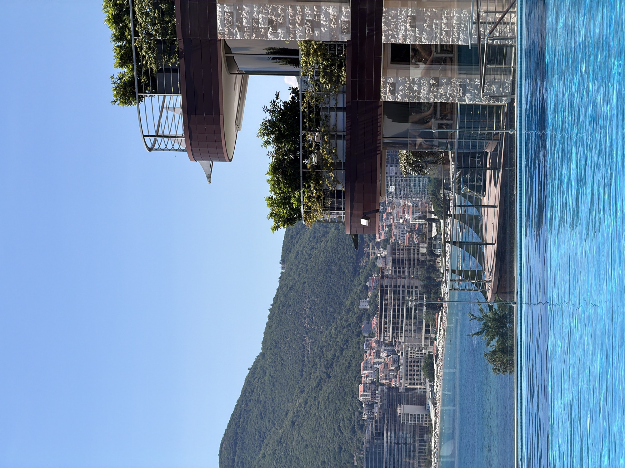 In Budva, Montenegro, a close-up view of a vibrant blue infinity pool dominates the foreground. Beyond the pool, a resort building with balconies adorned with lush greenery is visible, featuring modern architecture with stone accents and dark wood paneling. In the middle ground, the turquoise waters of the Adriatic Sea meet a coastal city characterized by a mix of modern high-rises and traditional red-roofed buildings. A verdant, tree-covered mountain slopes upward behind the city, under a clear blue sky. No people are discernible in the image.