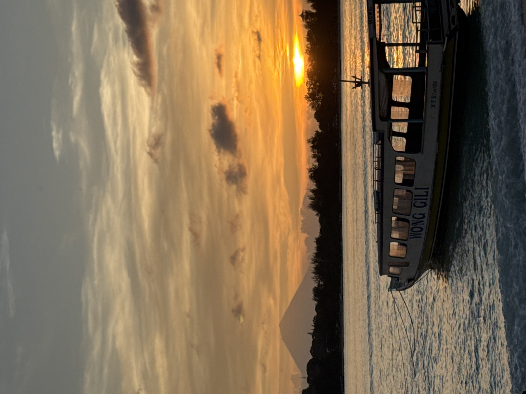 The image captures a boat with the name "WONG GILI" on its side in waters off the coast of Unknown, Indonesia. The photo is taken at what appears to be sunset, as the sky is filled with golden and orange hues and the sun's light is reflected on the water. There is some vegetation visible on the shore.