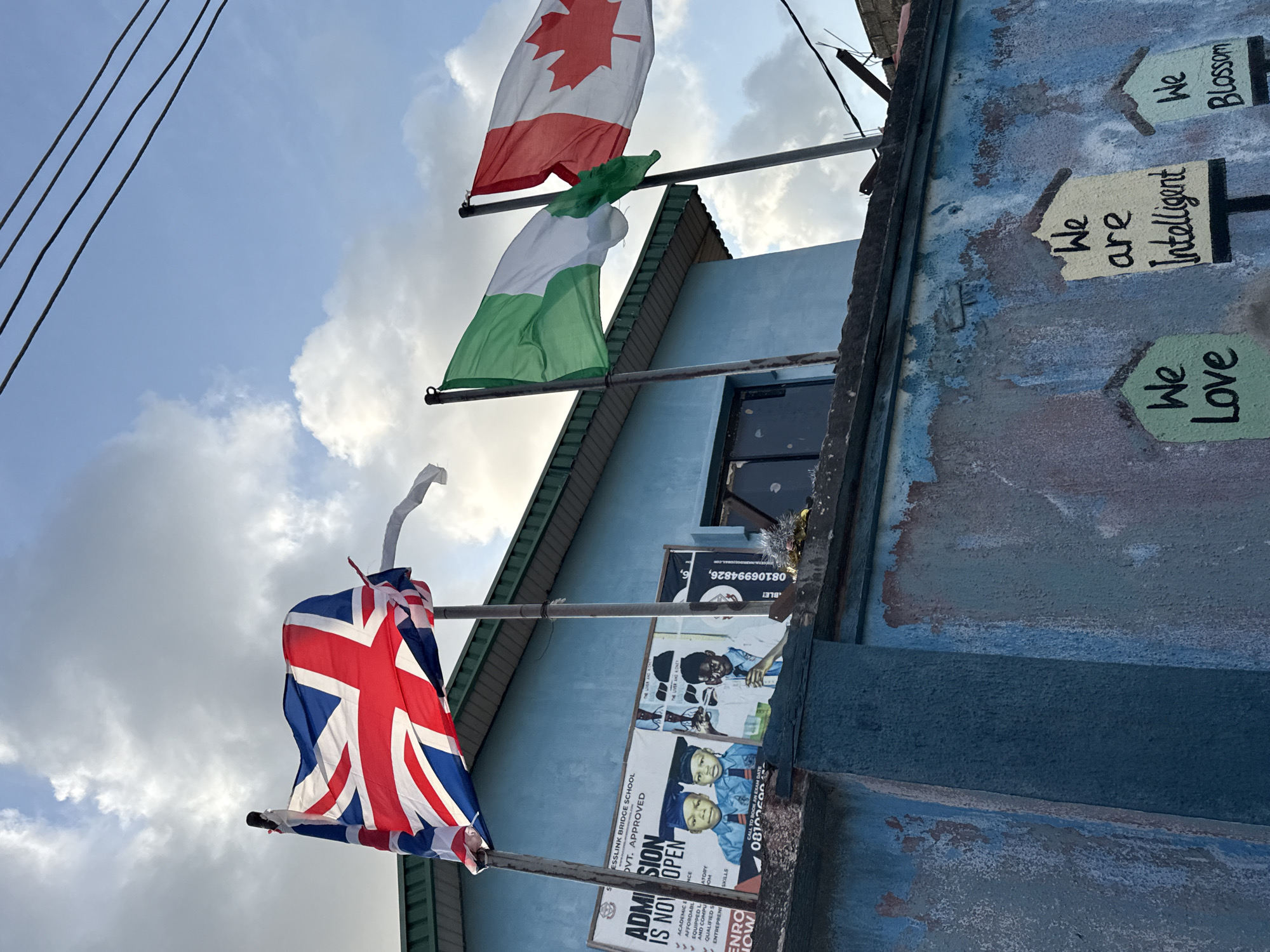 The image shows the exterior of Esslink Bridge School in Ado, Nigeria. Three flags are mounted on poles, in ascending order: the United Kingdom, Nigeria, and Canada. Below the flags are posters advertising school admission and containing phone numbers: 0810625000... , 08106994826... Educational mottos are painted on the building's facade: "We Love," "We are Intelligent," and "We Blossom." Power lines are visible against a cloudy sky. The building is blue and appears aged.