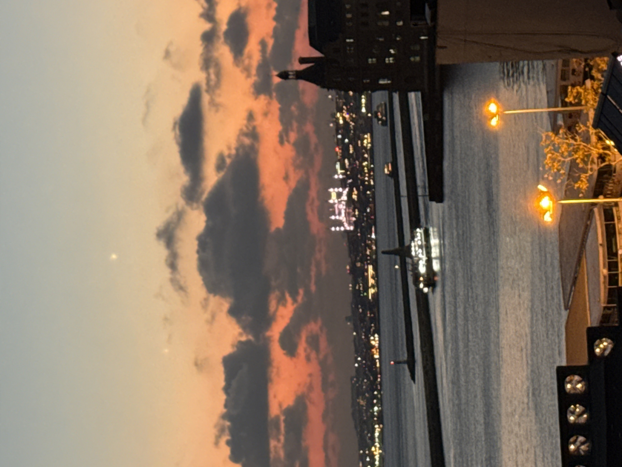 The image captures a twilight scene of the Istanbul skyline as viewed from Kadıköy, Turkey. In the foreground, two streetlights with bright orange light illuminate a portion of the waterfront. To the right, a portion of a building with illuminated windows is visible. Beyond this, the calm, dark water of the Bosphorus Strait separates Kadıköy from the European side of Istanbul. Two ferries or boats are seen navigating the water, with the lights of one clearly visible. A long, dark pier extends into the water, with a lighthouse at its end. In the distance, the skyline of the European side of Istanbul is dotted with illuminated buildings and lights, suggesting a city alive at dusk. A distinctive white building complex with numerous lights is prominent in the mid-distance. Above the city, the sky transitions from a soft orange hue at the horizon to a pale blue, with scattered dark clouds. Two bright celestial bodies, likely planets or stars, are visible in the upper portion of the sky. The overall impression is one of serene urban beauty at the transition from day to night.