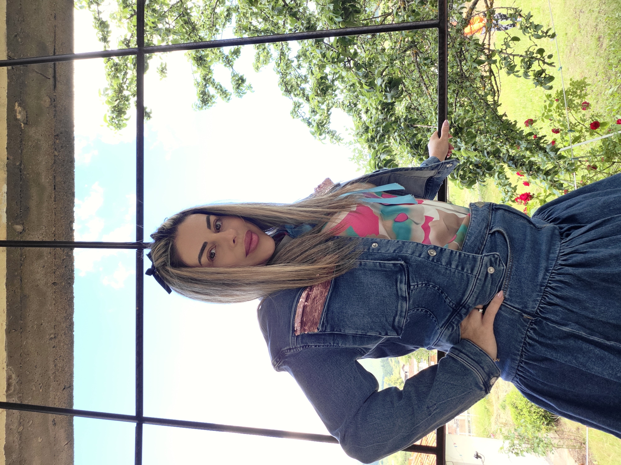 A woman stands in front of a window with a view of Ravnogor, Bulgaria. She is wearing a denim jacket and skirt set, a colorful top, and a black bow in her hair. Her left hand is on her hip, and her right hand rests on a window frame. The background shows a bright sky with scattered clouds, lush green trees, and some red roses. A building can be seen in the distance. The overall scene suggests a warm, sunny day outdoors.