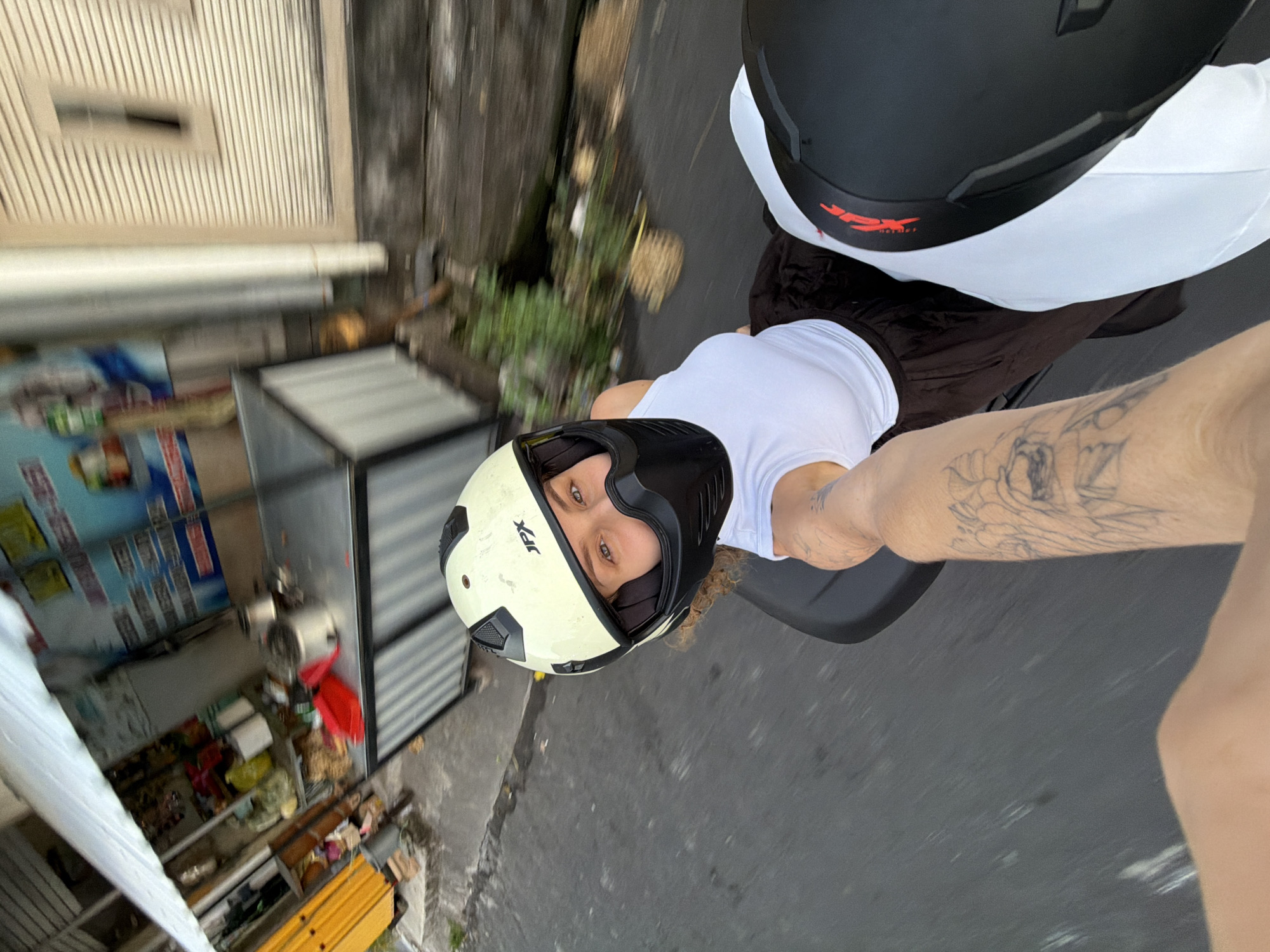 In North Kuta, Indonesia, a woman wearing a white tank top and a beige helmet with a black face mask is riding on the back of a motorcycle. The motorcycle is moving along a paved road, and the background shows a street scene with various stalls and buildings. A person in front, likely the driver, is holding out their arm, which is visible in the foreground with a tattoo on the arm. The image is taken from a dynamic, first-person perspective, suggesting the photo was captured while in motion.