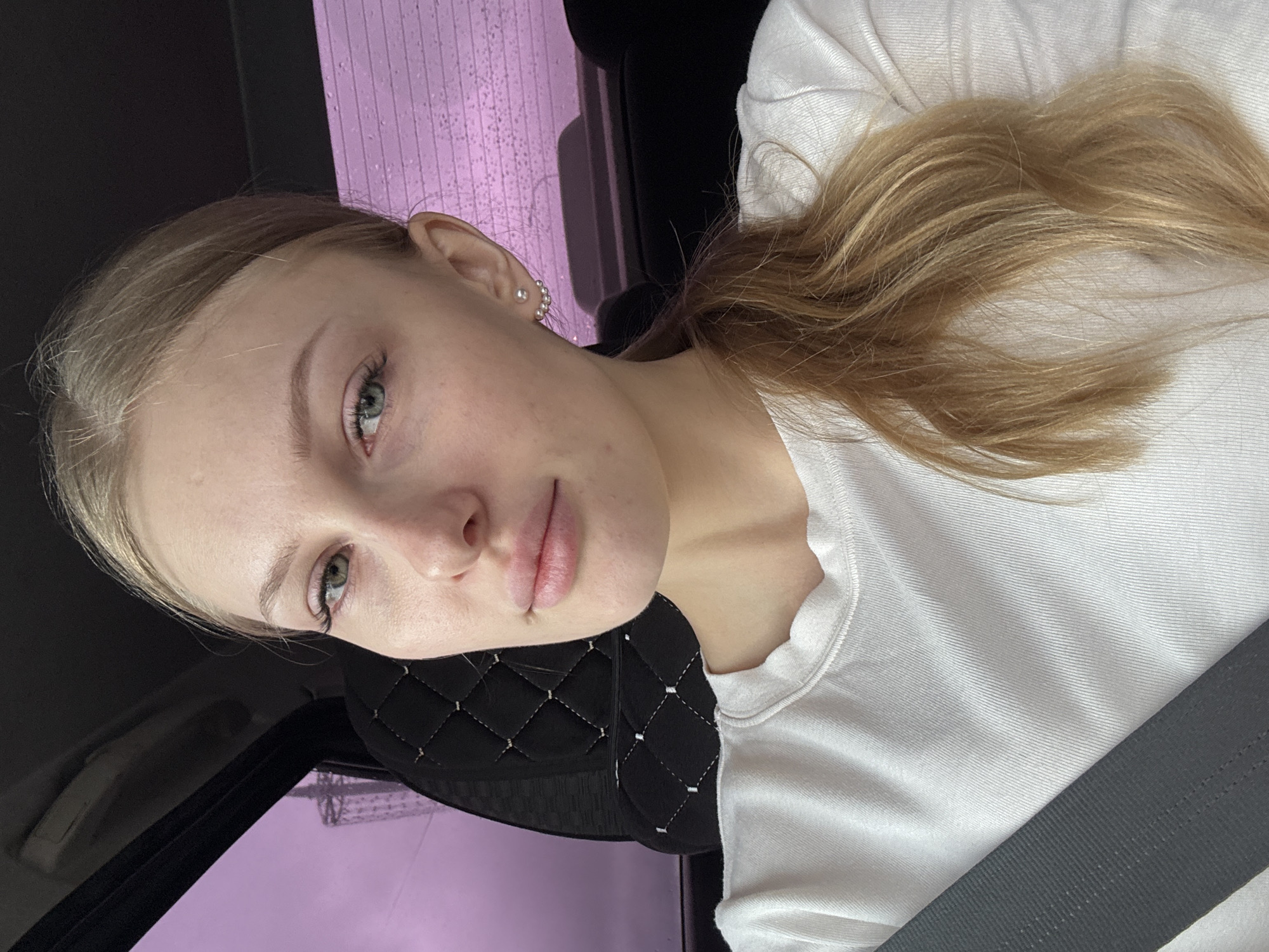 A young woman with blonde hair and green eyes is depicted in a close-up photograph. She is wearing a white shirt and a seatbelt, suggesting she is in a car. The background shows a pink wall and what appears to be a building. The location can be identified as the city of Perm, Russia, based on the appearance of the buildings and landscape. The woman has fair skin and is wearing earrings. The photo's angle suggests it might be a selfie taken inside the vehicle.
