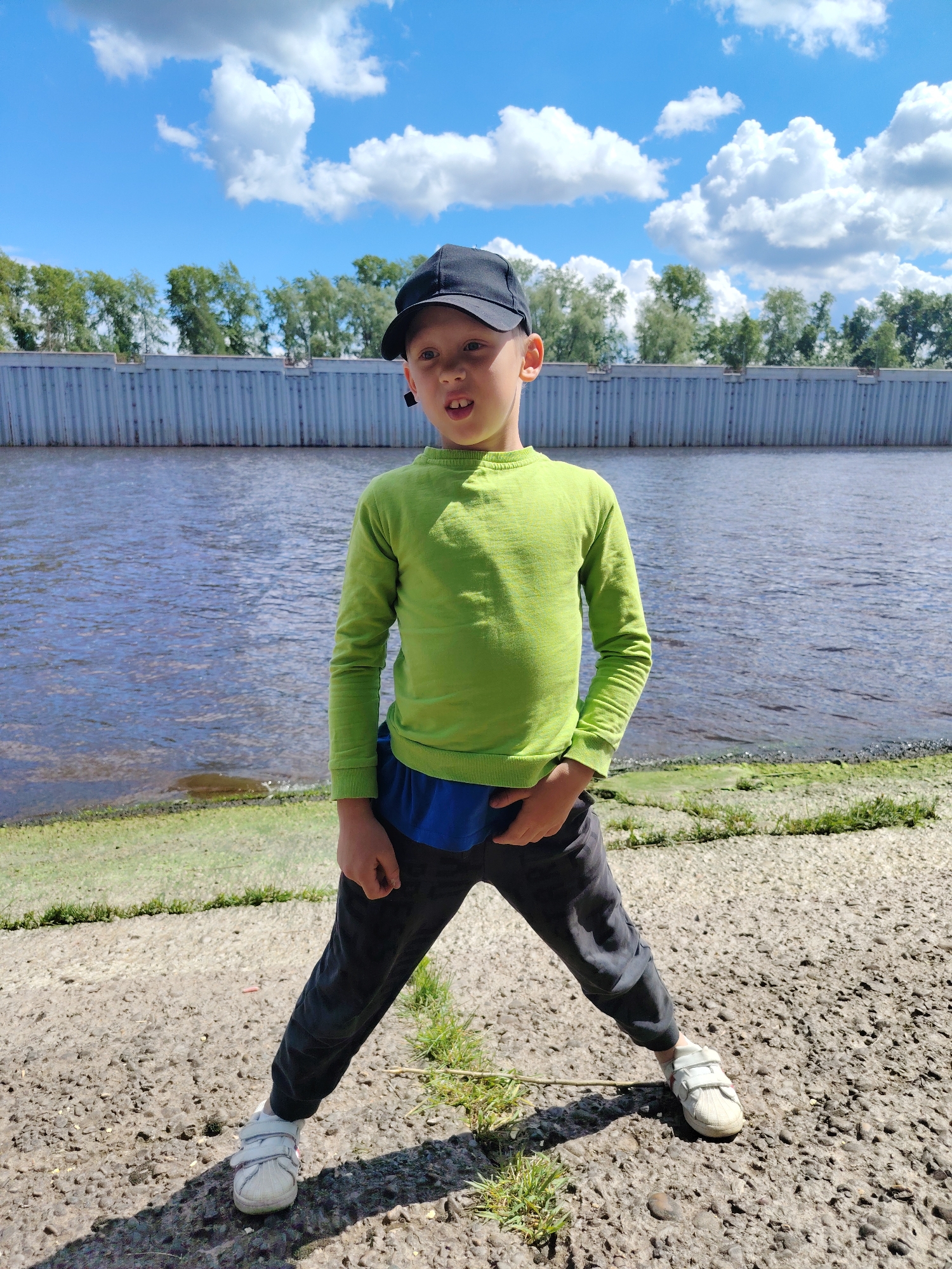A young boy stands outdoors, likely in a recreational area in Chaikovsky, Russia, based on known landmarks. He wears a green long-sleeved shirt, dark pants, white sneakers, and a black baseball cap. He is standing with his legs spread apart on a concrete surface, possibly a walkway or embankment. Behind him is a body of water, presumably the Kama River, with a vertical metal retaining wall along the shoreline. Green trees are visible in the background against a partly cloudy blue sky. The boy is looking slightly to his right with his mouth open. His left hand is in his pocket. The time of day appears to be daytime, with bright lighting.