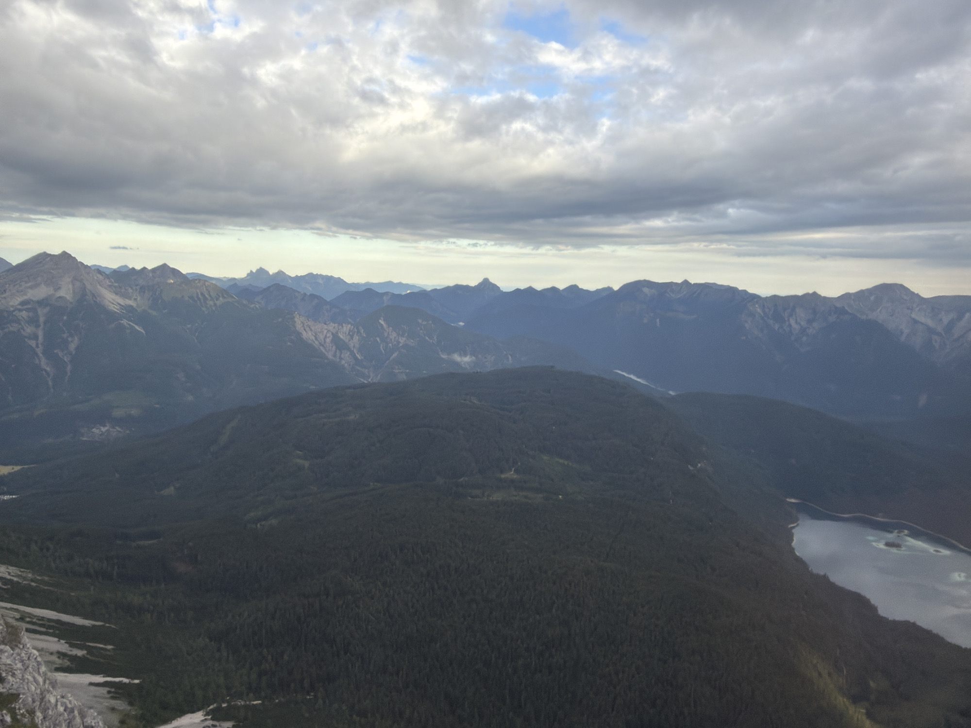 This is an aerial photograph capturing a vast mountainous landscape under a cloudy sky. The foreground is dominated by a dark, densely forested mountain slope that descends towards a body of water. The water appears to be a lake with a light blue hue, and a small island is visible within it. The middle ground features several layers of mountains, appearing progressively lighter and bluer with distance, indicating atmospheric perspective. The background is composed of rugged, snow-capped mountain peaks under a heavily clouded sky with patches of blue visible. The overall scene is natural and serene, with no human presence or man-made structures immediately apparent. The image appears to be taken from a high vantage point, possibly an aircraft or a very high mountain peak, looking out over the mountain range. The location is identified as Grainau, Germany, suggesting a view of the Bavarian Alps.