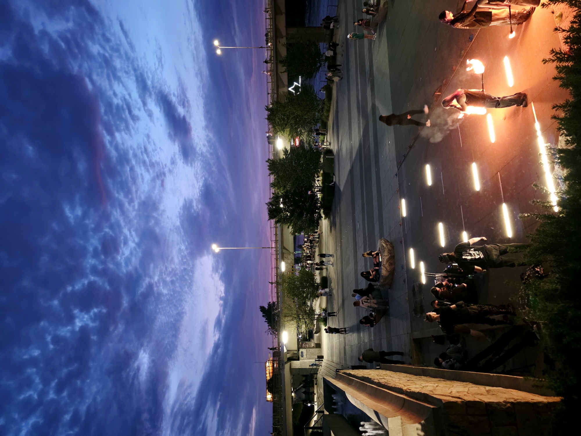 The image captures an evening scene in Warsaw, Poland, likely on the Vistula River embankment. The sky is a deep twilight blue, transitioning to purple and pink hues, with scattered clouds illuminated by the fading light. In the foreground, a group of people are gathered on a paved area, with some appearing to be performers engaged in an activity that involves fire.
One individual, with long hair, is holding two flaming props, creating a dramatic fiery effect. Another person beside them, wearing a dark shirt, is also holding a flaming prop. Several other people are visible in the background, some walking, some sitting, and some standing around the performers. The area is illuminated by overhead streetlights and the bright glow from the fire.
Further back, a pedestrian bridge spans across the scene, with lights visible on its structure and traffic moving across it. Trees are planted along the embankment, and a fountain with water jets is partially visible on the left side of the image. The overall atmosphere suggests an outdoor public event or performance taking place as dusk settles over the city.