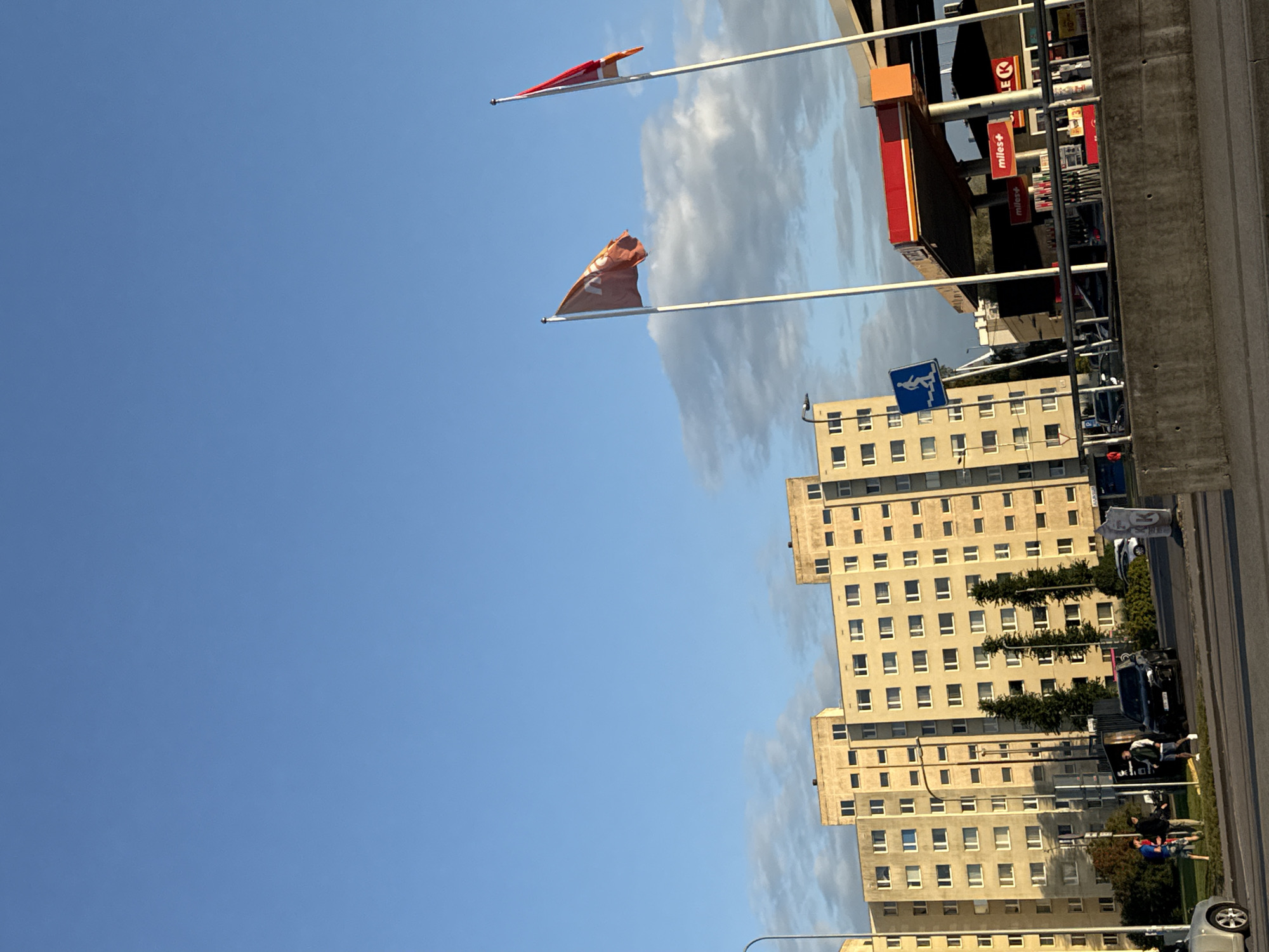 The image depicts a scene in Tallinn, Estonia, under a clear blue sky with scattered clouds. In the foreground, a concrete barrier runs horizontally. Beyond the barrier, a street is visible with cars, including a black SUV and a white sedan. A gas station with a red and orange canopy is prominent, featuring signs for "Miles+" and a red circular logo with a "K". Two flags fly from poles near the gas station; one is orange with an emblem, and the other is red with a white design. In the background, a tall, light-colored apartment building with numerous windows stretches across the frame. Several trees stand between the apartment building and the street. Three individuals are standing near the apartment building, appearing to be conversing. One is wearing a blue jacket, another a dark jacket, and the third a light-colored jacket. A blue sign with a white silhouette of a person climbing stairs is mounted on a pole.