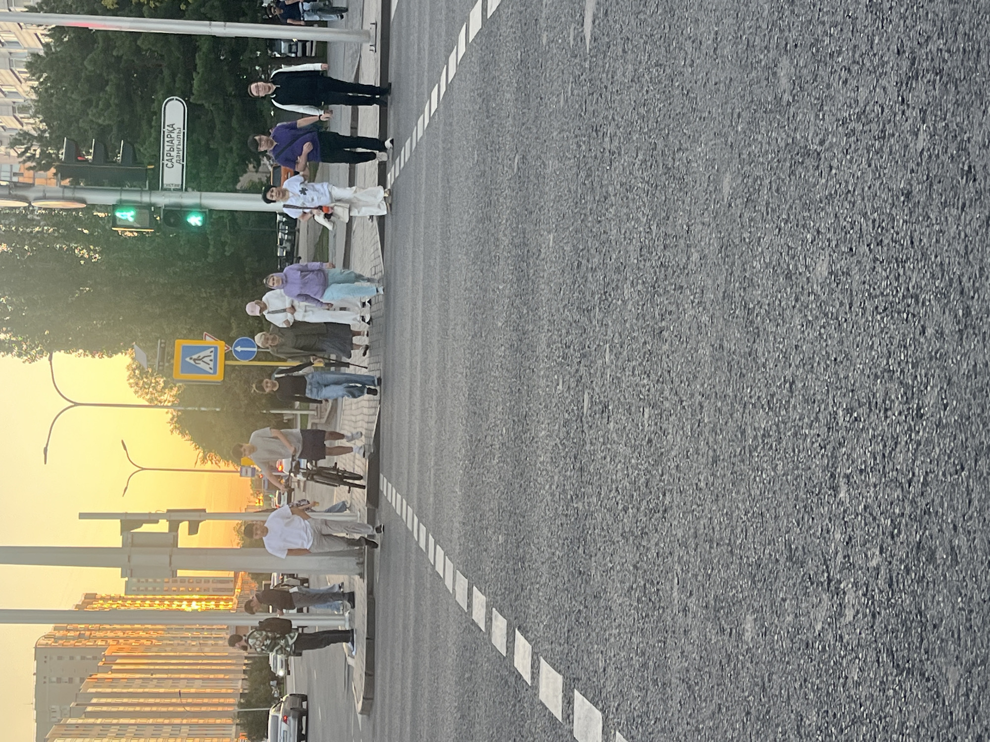 The image depicts a street scene in Astana, Kazakhstan, at sunset. A group of pedestrians is crossing a wide asphalt road at a marked crosswalk. The sun is low in the sky, casting a warm, orange glow over the scene, illuminating the tall, modern buildings on the left side of the street. Traffic lights, showing green for pedestrian crossing, are visible on a pole, along with a street sign indicating "Saryarka Avenue."
In the foreground, the crosswalk is marked with white dashed lines. The people crossing the street are diverse in age and gender. One individual is walking a bicycle. The group includes individuals in casual attire such as t-shirts, jeans, and hoodies. On the sidewalk to the left, several other people are standing, some appearing to be waiting or observing. Cars are visible on the road in the background, and lush green trees line the right side of the street. The overall atmosphere is one of urban life in the evening.