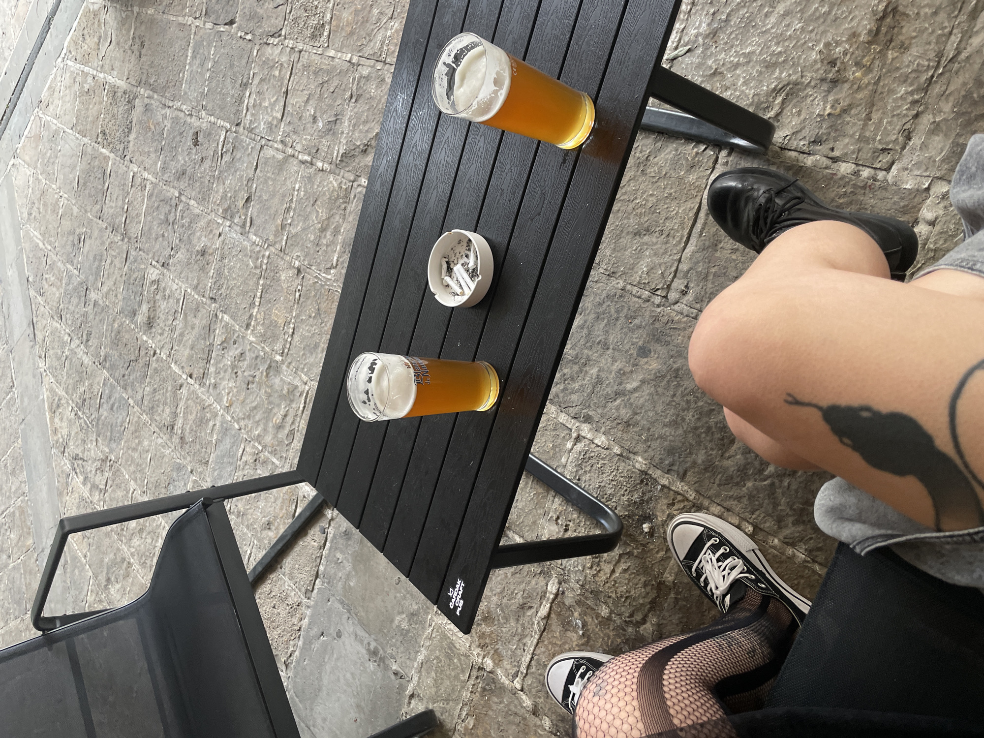 The image presents an elevated view of an outdoor table scene in Budva, Montenegro. A black, slatted table is the focal point, hosting two glasses of beer and an ashtray filled with cigarette butts. The table legs are black metal. A black chair, also with metal legs, is partially visible on the left.
To the right of the table, a person is visible, with a portion of their legs and feet in the frame. They are wearing black and white Converse sneakers, one foot with fishnet stockings. A snake tattoo is visible on their thigh. The second person is wearing black leather boots. The paving stones of the outdoor area are predominantly grey.
