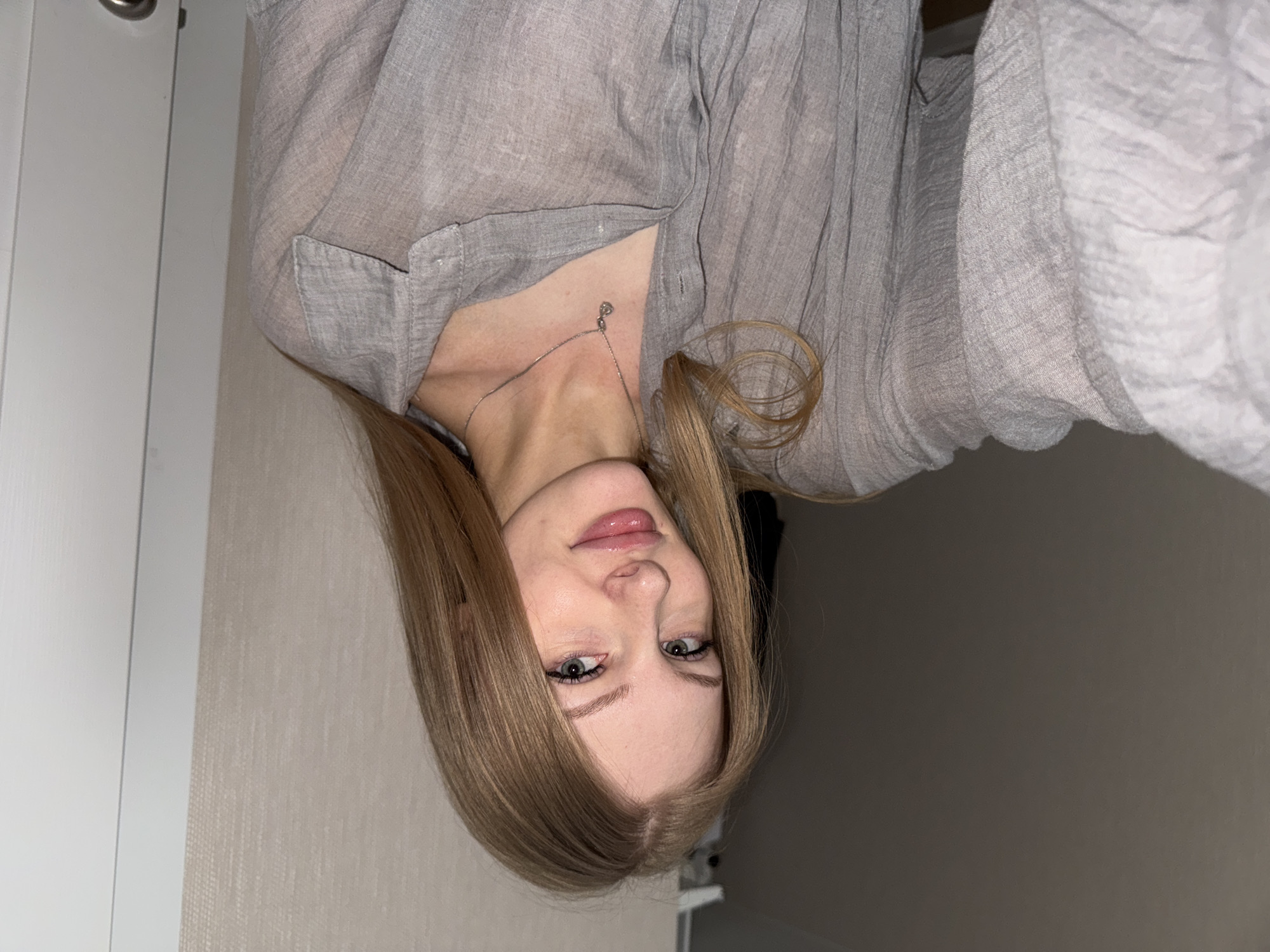In a medium shot, a young woman with long blonde hair and green eyes looks directly at the camera. She is wearing a light gray, loosely-buttoned shirt and a silver necklace with a small pendant. The background consists of a neutral-colored wall with a textured pattern and a white doorframe to her right. The lighting suggests an indoor environment, possibly a home. The image was taken in Perm, Russia.