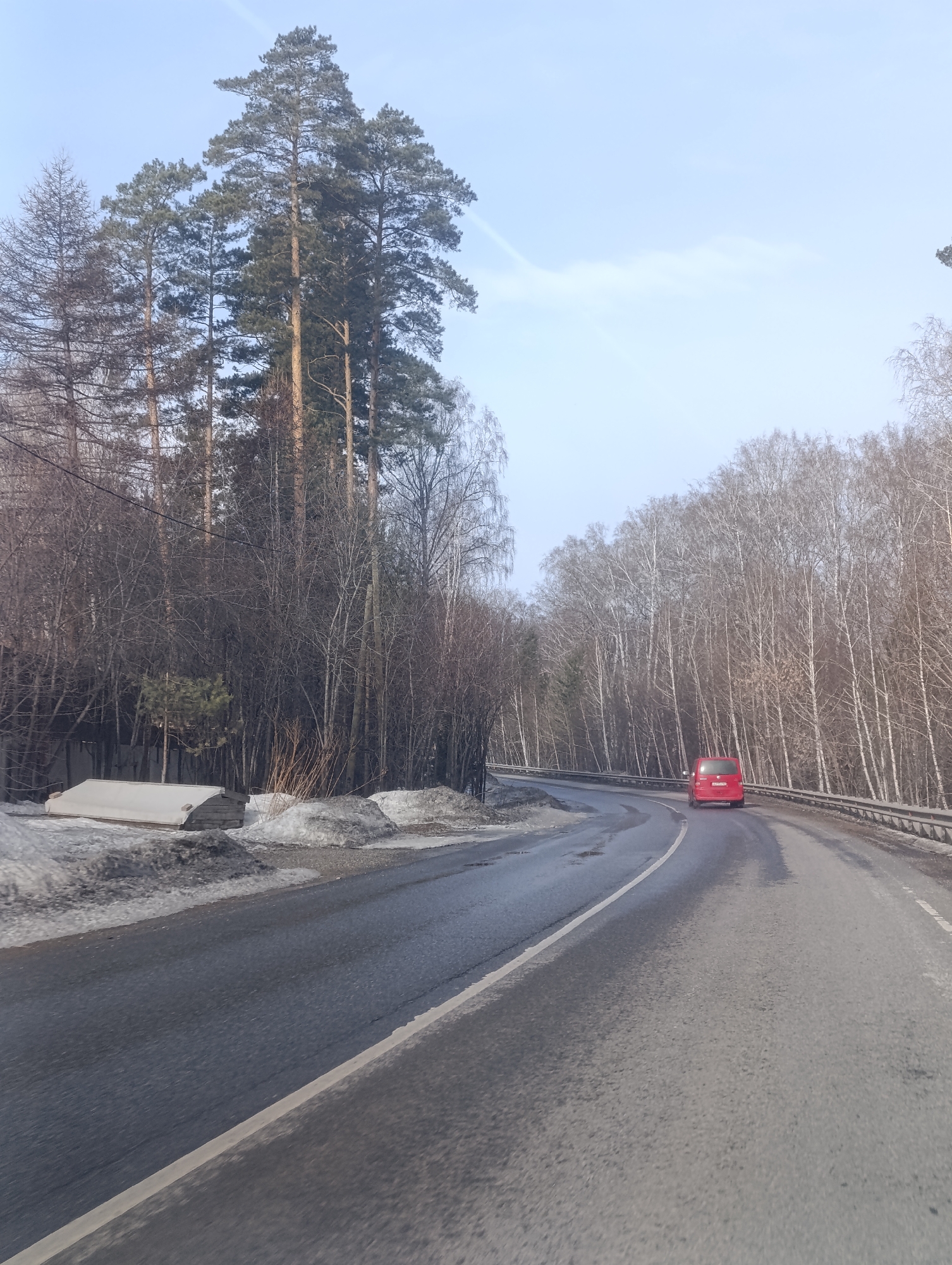 The image captures a road winding through a forested area. A red car is traveling on the road away from the camera. The road appears wet, with melting snow present on the side. The trees are mostly bare, indicating either winter or early spring. On the left of the road, there are some structures covered in snow. The sky is pale blue. The location is Krasnoyarsk, Russia.