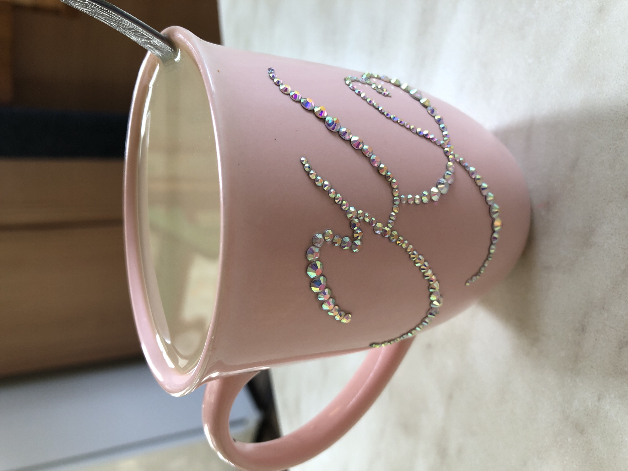 The image depicts a close-up, elevated view of a single light pink ceramic mug resting on a light-colored, possibly speckled, countertop or table surface in an indoor setting within the city of Noviy, Russia. The mug features a handle on its left side and is adorned with numerous small, iridescent, multi-faceted rhinestones arranged to form an elaborate cursive design on its exterior, resembling the letter "K" or a stylized initial. The mug contains a light-colored liquid beverage, consistent with coffee or tea with milk, and a silver-colored metal spoon with an ornate handle is partially submerged within the liquid, angled towards the upper right of the mug. The background is out of focus, showing blurred segments of a light blue-grey wall or object on the left and a darker brown, possibly wooden, surface on the right. No individuals are visible.