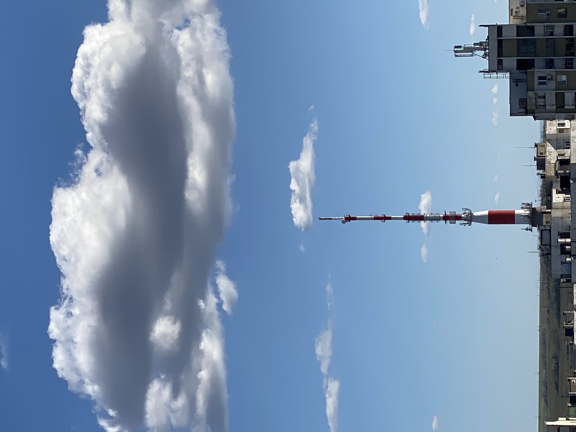 The image depicts a view in Pleven, Bulgaria, featuring a tall radio tower with red and white sections against a blue sky with scattered clouds. A large, dense cumulonimbus cloud is prominently positioned on the left. To the right of the tower are high-rise apartment buildings. A distant, green landscape stretches horizontally in the lower portion of the image. The image has been rotated 90 degrees counterclockwise.