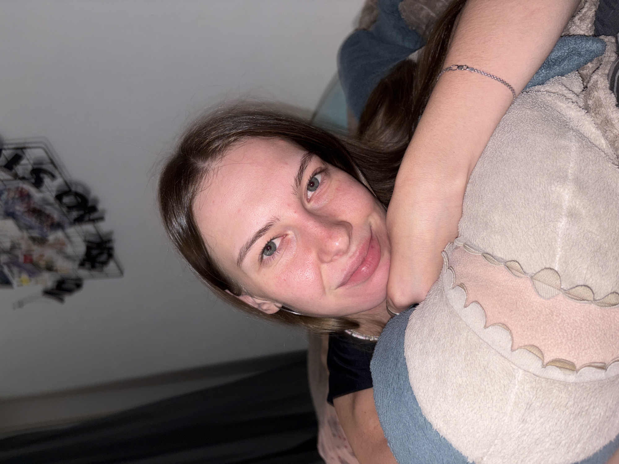A young adult female with light brown hair is lying on a bed, resting her head on her arm, which is propped on a pillow. She is looking at the camera with a slight smile. She is wearing a black t-shirt and a silver bracelet on her left wrist. The background shows a wall with some decorations and a curtain. The room appears to be a bedroom. There is no visible indication of the city of Perm, Russia.