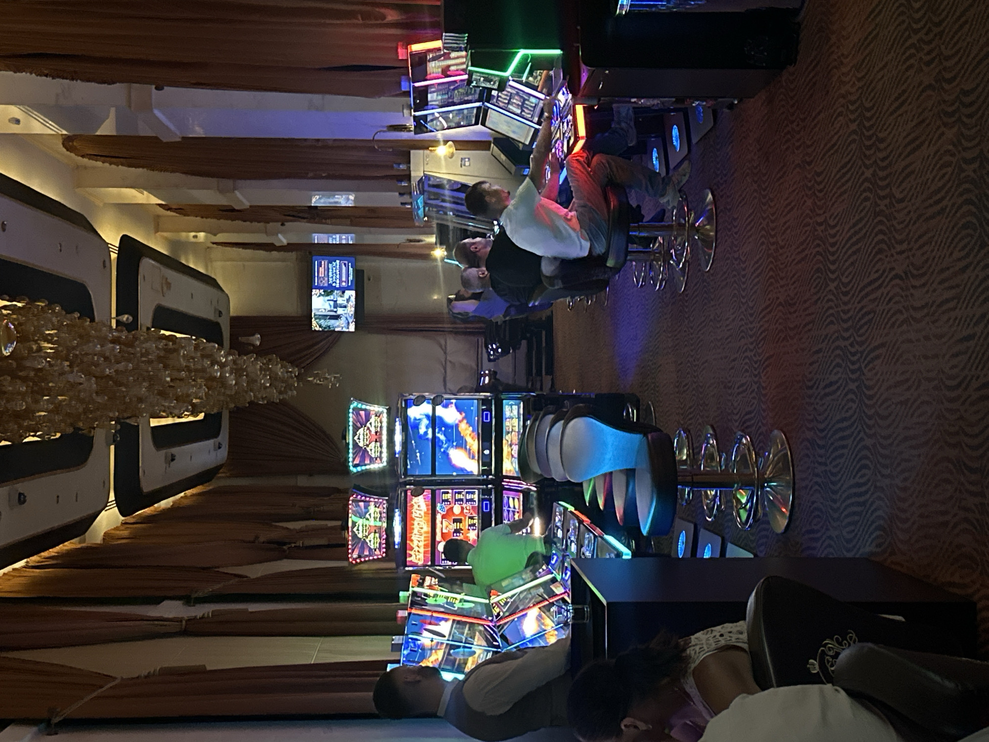 The image depicts the interior of a casino, likely located in Estosadok, Russia, given the context. The scene is dimly lit, with the primary illumination coming from the vibrant lights of numerous slot machines and overhead chandeliers. Several people are visible, some playing the slot machines and others seated on bar stools. The slot machines are arranged in rows, with their colorful screens displaying various graphics and animations. The flooring is carpeted with a patterned design, and the walls are adorned with heavy, floor-to-ceiling drapes. The overall atmosphere suggests a typical casino environment, focused on gambling entertainment.