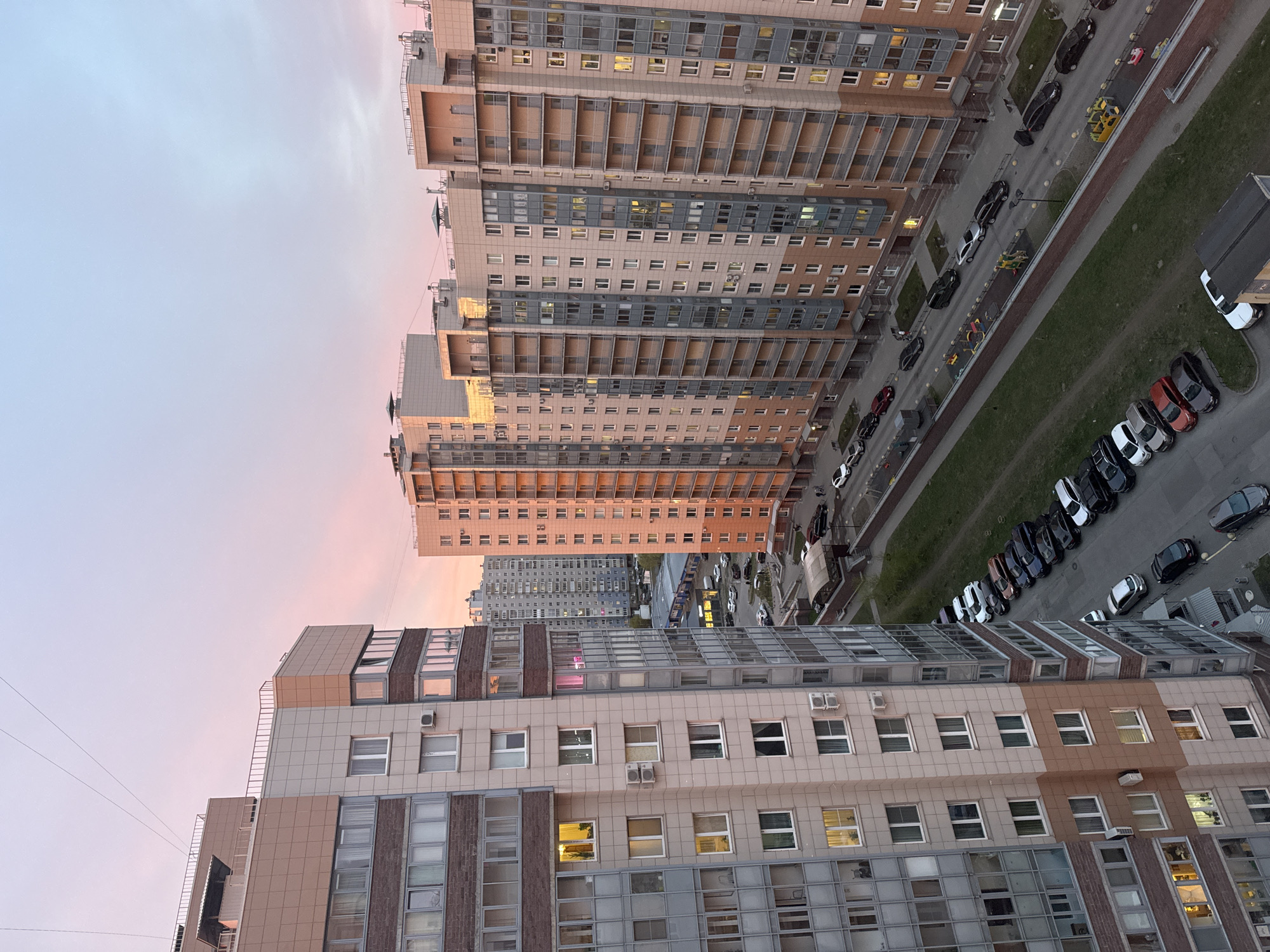 The image shows a view from a high vantage point, likely an upper floor of a building, overlooking an urban area at dusk or dawn, based on the pink hue in the sky. The foreground is dominated by the side of a multi-story building with a facade featuring a mix of beige, tan, and grey tiling, with rows of windows and what appear to be enclosed balconies or sunrooms on the upper floors. To the right of the foreground building, there is a street lined with parked cars in various shades of black, white, and red. A strip of greenery runs alongside the street. In the distance, another taller multi-story building with a grey and beige exterior and rows of balconies is visible, partially lit by the setting or rising sun. Further in the background, a smaller building can be seen amidst the urban landscape. The overall impression is of a residential or mixed-use area with a relatively high density of buildings.