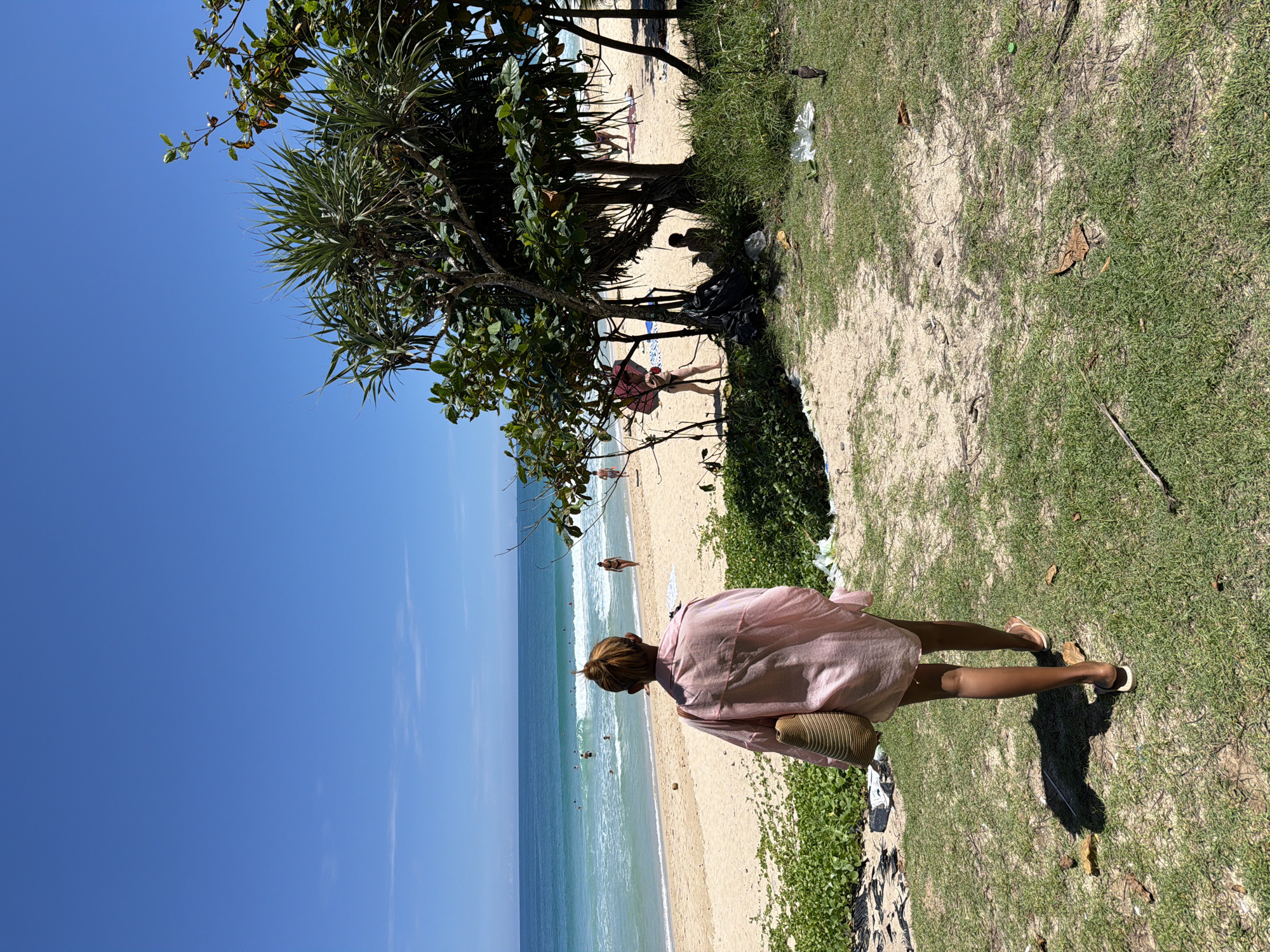 In the city of Choeng Thale, Thailand, a woman with her back to the camera walks on a grassy area adjacent to a sandy beach. She is wearing a pink striped collared shirt and carrying a woven bag over her left shoulder. She is wearing sandals. The beach is populated with several people, some swimming in the turquoise ocean, others walking along the shore. Lush green vegetation borders the sandy beach, and several trees with spiky leaves are visible, casting shadows on the sand. The sky is a clear, bright blue. The environment is a tropical beach scene on a sunny day.