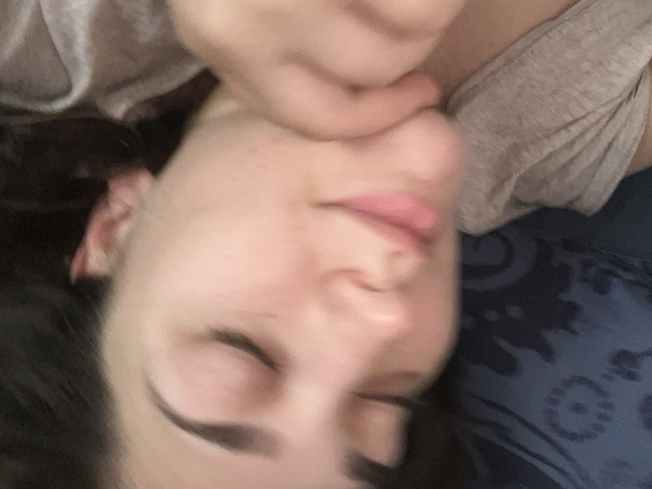 The image captures a close-up of an individual, identified as likely an adult female, resting in an indoor setting within Ancenis, France. Her eyes are closed, and a subtle smile is present on her lips, suggesting a state of sleep or deep relaxation. The individual possesses fair skin, with visible dark eyebrows and dark hair partially obscuring the top of the frame. A hand, presumably her own, supports or rests near her jawline and upper neck on the right side. She is dressed in a light grey short-sleeved garment, possibly a t-shirt. Her head is supported by a dark blue fabric object, likely a pillow, adorned with a lighter, indistinct patterned design. The scene is illuminated by soft, ambient lighting.