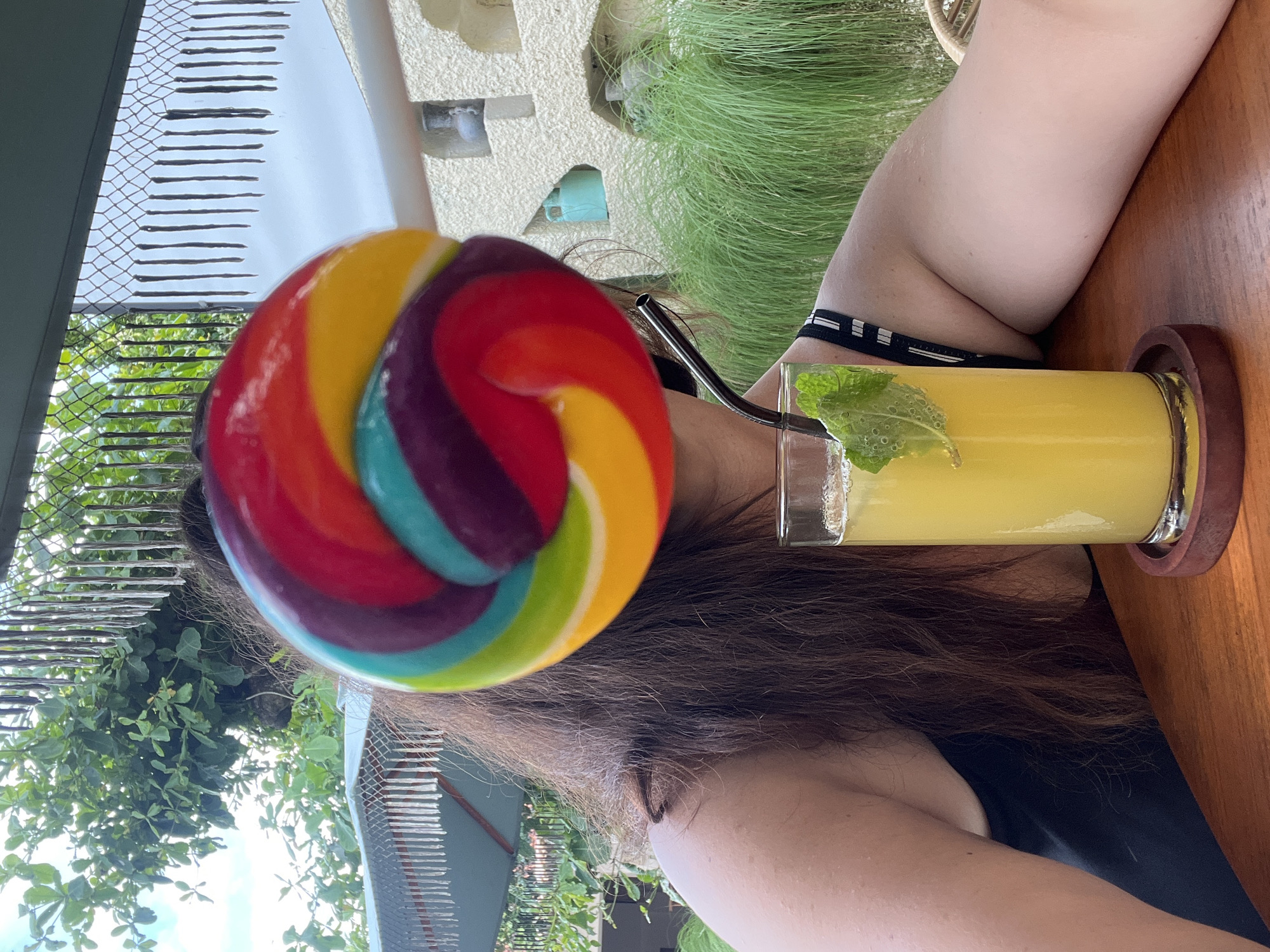 A person is holding a colorful lollipop in front of their face, obscuring their features. They are seated at a wooden table, with a tall glass of yellow liquid, a straw, and mint leaves in front of them. The person is wearing a black top. The background suggests an outdoor setting, possibly a cafe or restaurant, with lush green foliage, a white stucco wall with decorative cutouts, and some hanging decorative elements. The scene appears to be in a tropical environment.