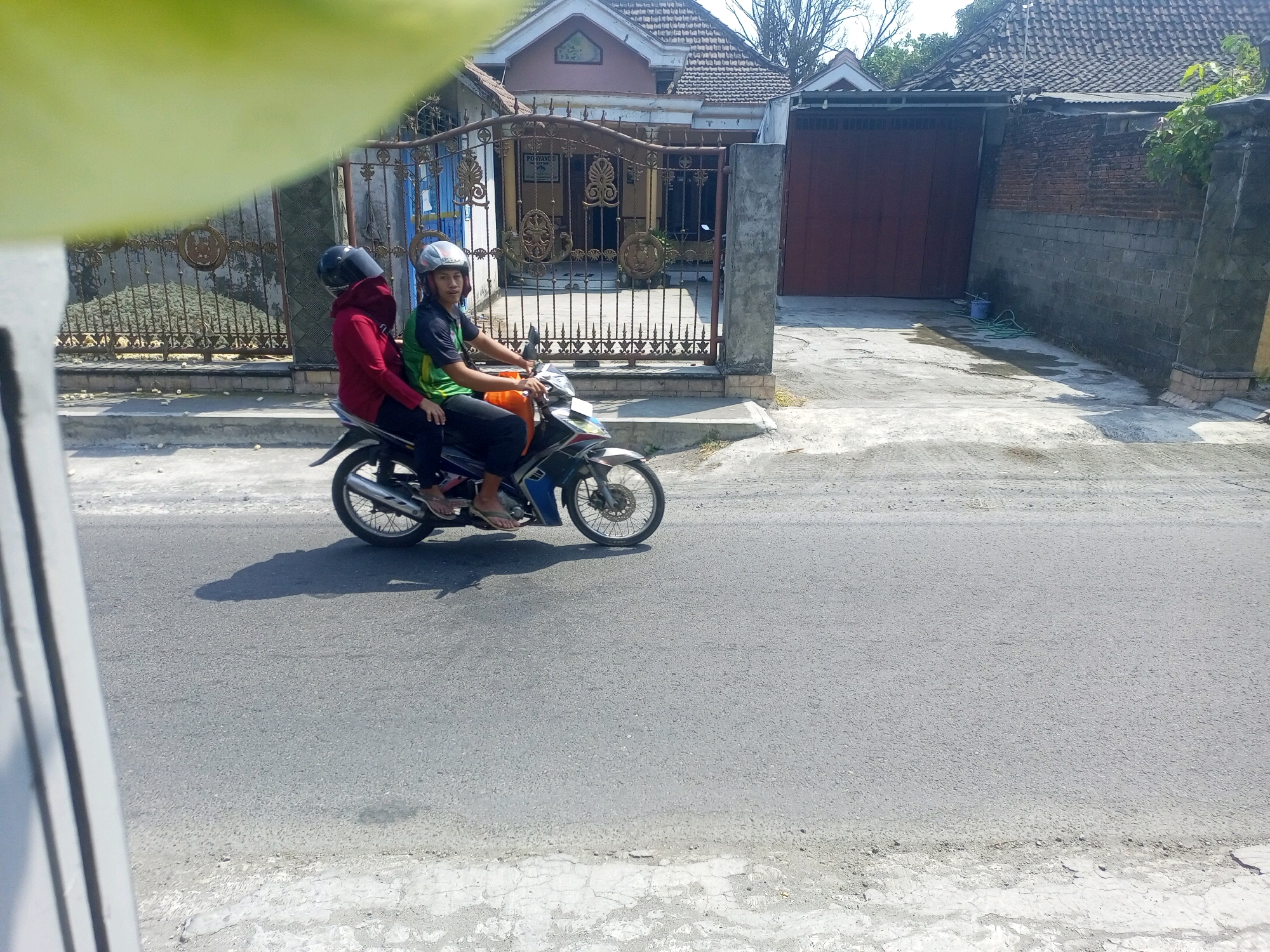 Two individuals are riding on a motorcycle on a paved road in what appears to be a residential area. The rider in front is wearing a green and black shirt and a silver helmet. The passenger behind is wearing a red top and a black helmet. They are both facing forward, suggesting they are traveling. The motorcycle is a light blue and silver model. In the background, a house with a tiled roof and a decorative metal gate is visible. A large brown gate is also present on the right side of the image. The road surface is asphalt, and there is some vegetation on the right side of the scene. The lighting indicates it is daytime with bright sunlight casting shadows. The overall setting suggests a street in Ngasem, Indonesia.