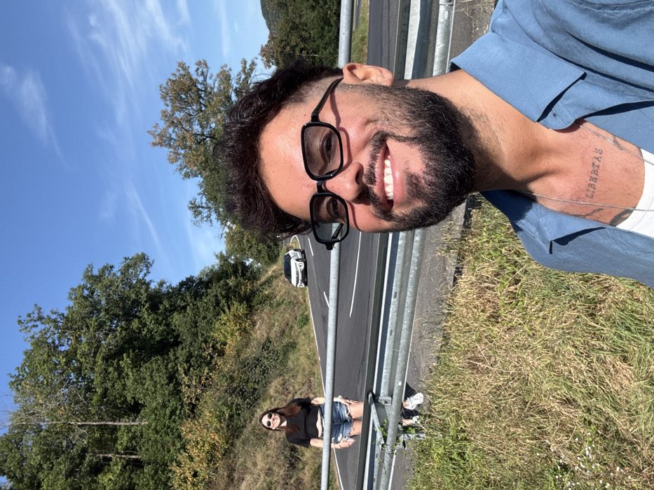 The image depicts a sunny outdoor scene on a rural road within the Cochem region of Germany. In the immediate foreground, slightly to the right, a man with dark hair, a beard, and dark-framed sunglasses smiles broadly at the camera, taking a selfie. He is wearing a light blue open-collared shirt over a white undershirt, revealing a silver chain necklace and a "LIBERTAS" tattoo on his upper chest.
Behind him, extending across the mid-ground, is a silver-colored metal guardrail. To the left of the frame and behind this guardrail, a woman stands on the roadside. She has long, dark hair, wears sunglasses, a black cropped t-shirt, light blue denim shorts, and white sneakers. She looks towards the camera with a slight smile.
Beyond the guardrail, a two-lane asphalt road curves into the distance. A white SUV or crossover vehicle is visible on this road in the background, heading away from the camera. The road is flanked by steep, vegetated hillsides. The foreground features dry, sparse grass, while the hillsides are covered in dense deciduous and coniferous trees, with some leaves exhibiting early autumnal colors. The sky above is clear blue with minimal wispy clouds.