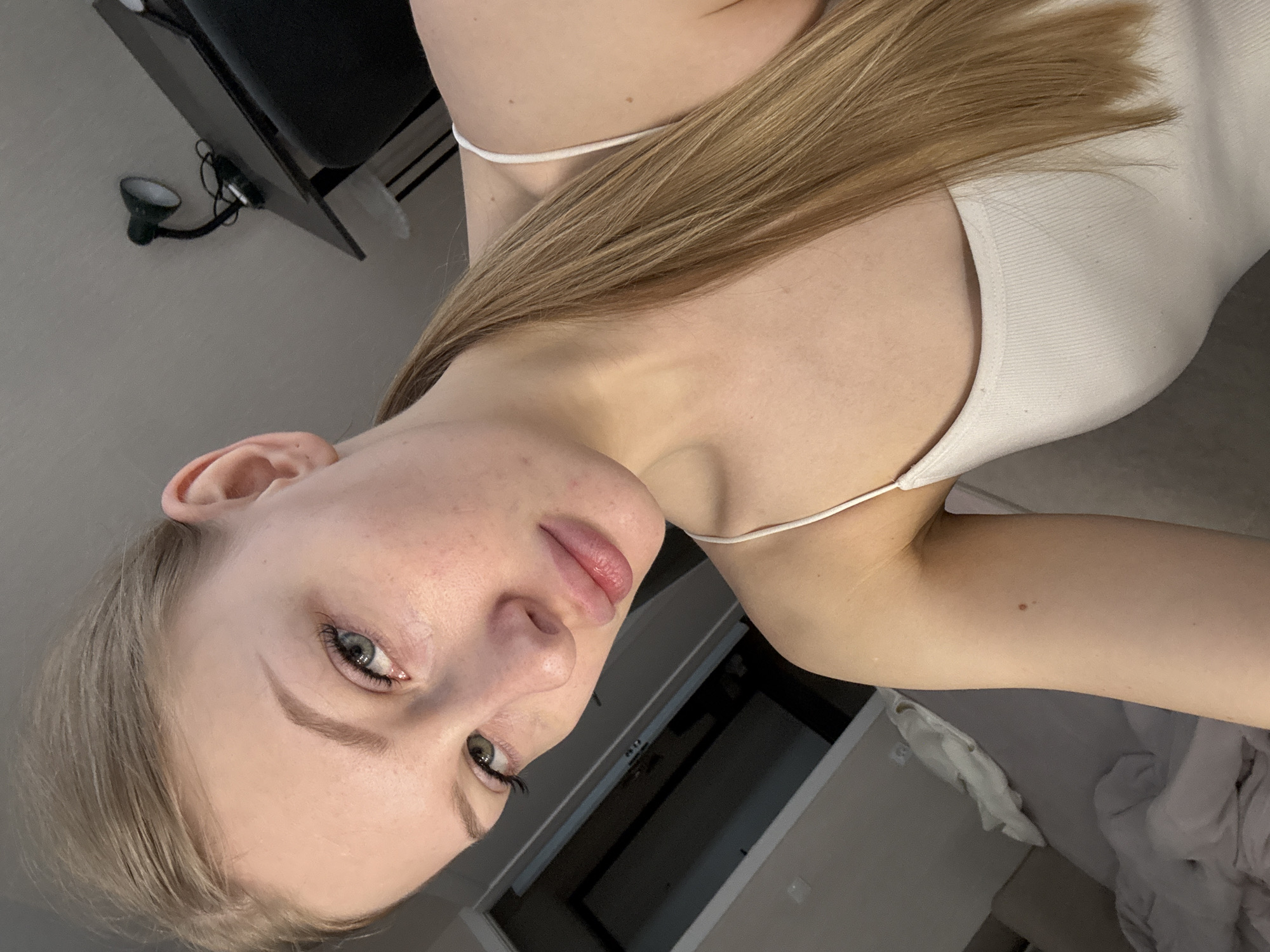A young woman with fair skin, blonde hair, and blue eyes is posing for a selfie. She is wearing a white tank top with thin straps. The background includes a wall with a lamp, a dark object that appears to be a piece of furniture, and a partially visible bed with white bedding. There is a doorway or archway visible in the background. The location is undetermined.