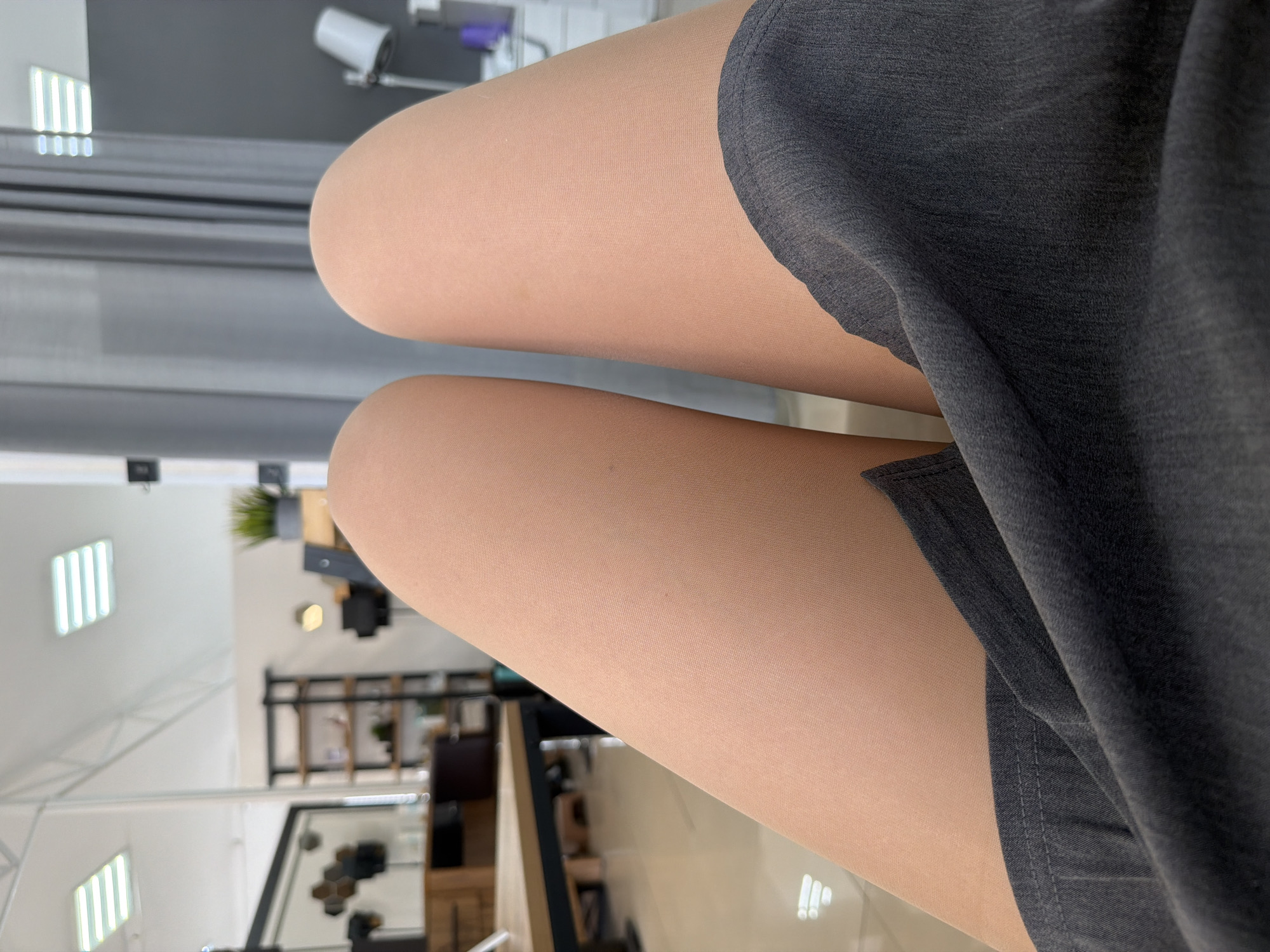 The image shows a close-up view of a person's legs wearing flesh-colored tights, with a dark grey garment partially covering the upper thighs. The background is a blurred interior scene, which includes elements like lighting fixtures, shelves, and furniture. The environment appears to be a shop or office setting. The location is Chaikovsky, Russia.