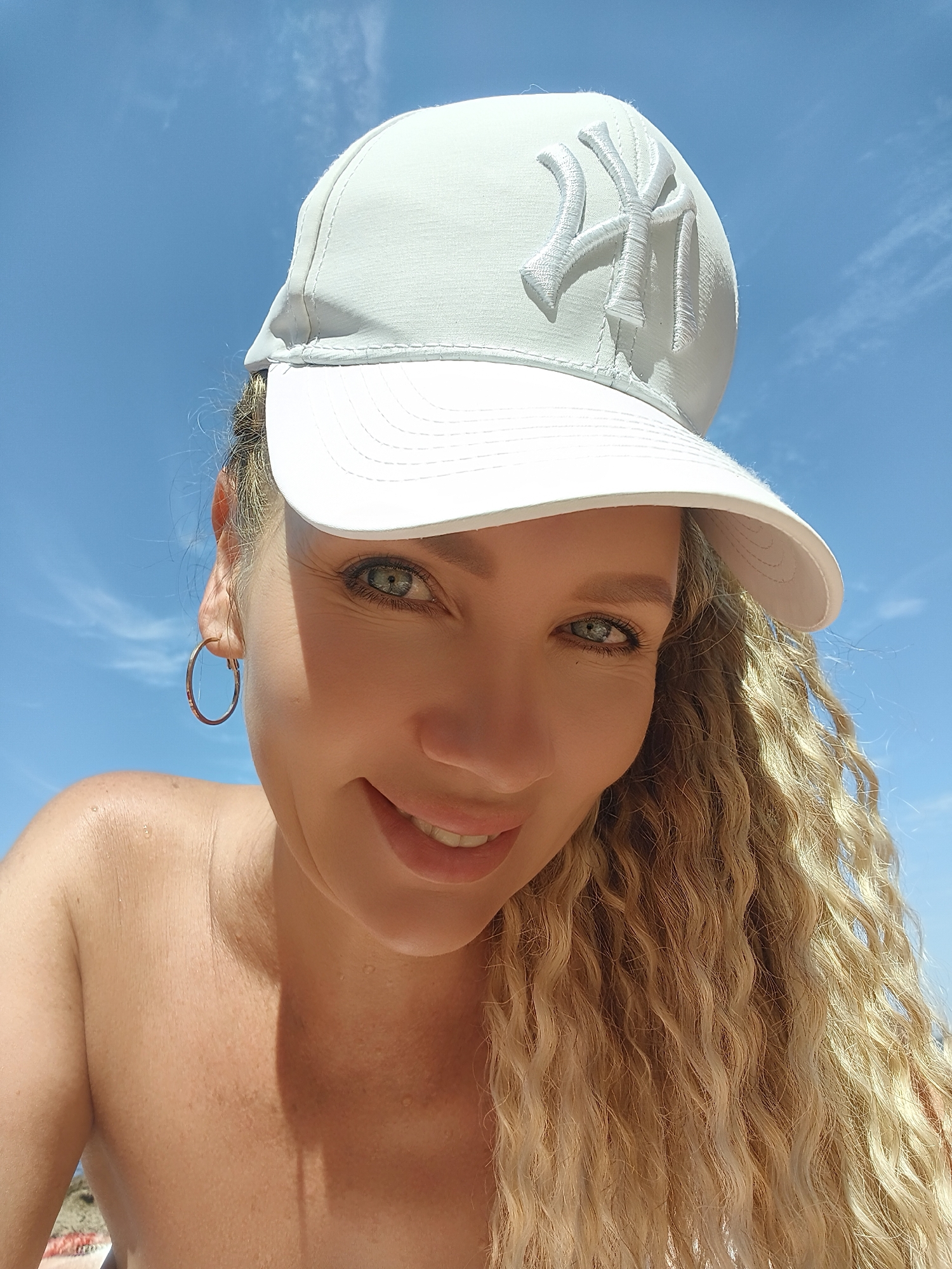 A woman with curly blonde hair and blue eyes is wearing a light gray baseball cap with a New York Yankees logo. She is also wearing a hoop earring. The woman is outdoors, with a blue sky and white clouds in the background. Her shoulders and chest are visible, and she appears to be at a beach or other outdoor recreational area. There are small water droplets on her skin, suggesting she may have been swimming or it is humid. The image is a close-up selfie. There is no information to suggest the location is in Chaikovsky, Russia.