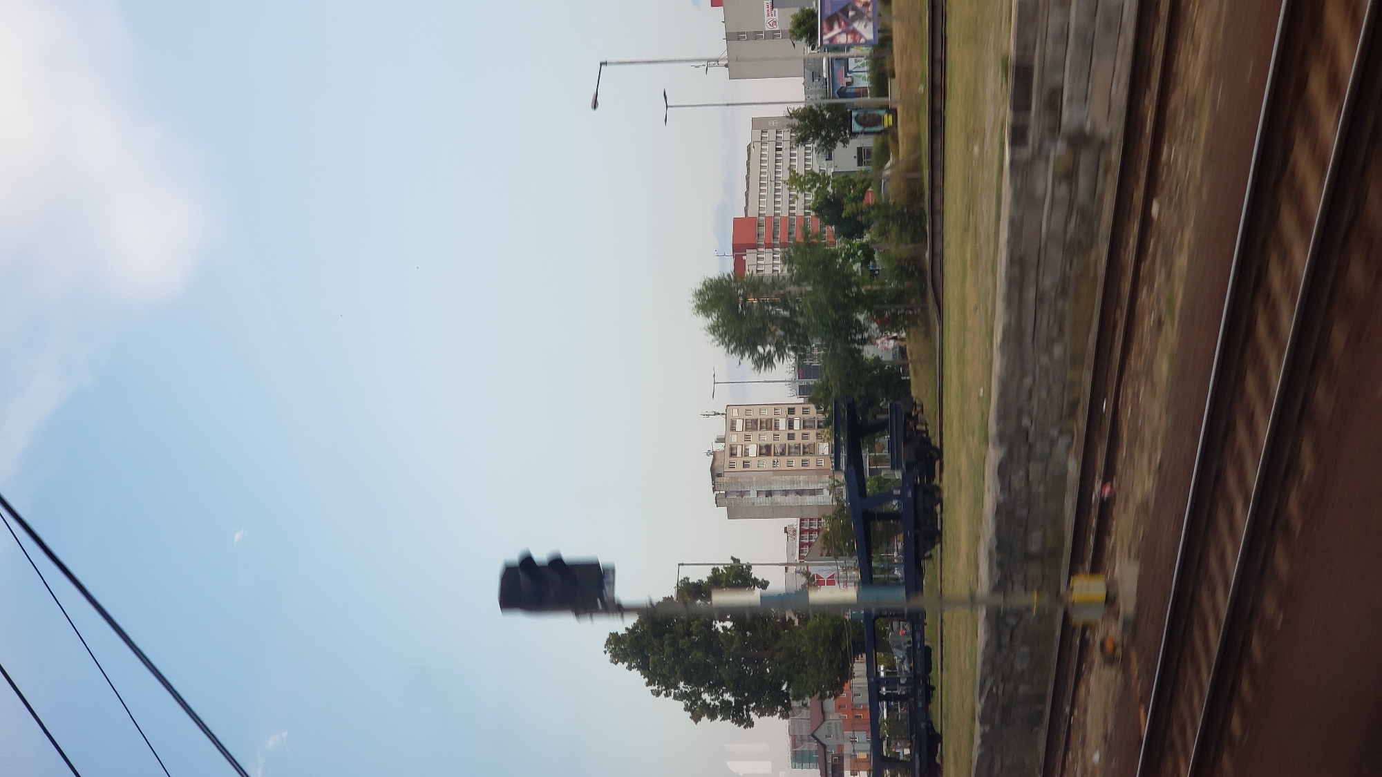 This is a vertical, eye-level, outdoor photograph taken from a moving vehicle, likely a train, looking towards a cityscape. The sky is clear and light blue with some wispy white clouds. In the foreground, several train tracks are visible, running horizontally across the bottom of the frame. The tracks are somewhat blurred due to motion.
Beyond the tracks, there is a grassy area with a low, stone wall. A tall, dark traffic signal is prominent on the left side of the image, slightly out of focus. Behind the traffic signal, a large, green tree stands tall. Further in the background, several multi-story buildings line the horizon. One building is a light-colored, square apartment block with many windows. Another building to its right has a red and white facade. Other buildings are visible further in the distance, some appearing to be commercial structures with signage. There are also streetlights and what appear to be cell towers on some of the buildings.
A dark-colored train car, likely a flatbed, is situated on tracks in front of the buildings. Some advertising posters are visible on the right side of the image, attached to what seem to be kiosks or bus stops. The overall impression is of a view from a train entering or passing through an urban or suburban area of Poprad, Slovakia, with a mix of residential and commercial buildings and infrastructure. No people are clearly discernible in the image.