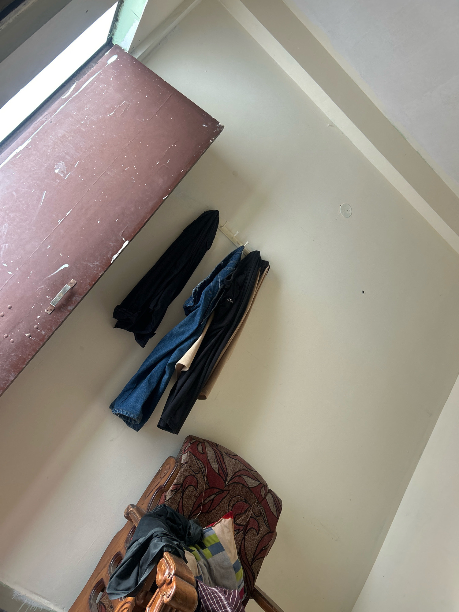 An interior scene, shot from a low angle looking upwards, depicts a cluttered corner of a room. To the left, a portion of a large, dark reddish-brown door, possibly a closet or room door, is visible, showing signs of wear and a metal handle. Above the door, a window with a green frame is partially open, letting in bright light. Against the off-white wall, several items of clothing are hung on a white rack: a black garment, blue jeans, and another black garment. Below the hanging clothes, a wooden chair with an ornate carved frame and patterned upholstery is partially visible, with more dark clothing draped over its back and seat. The overall impression is one of a lived-in, somewhat disorganized space. The scene is not specifically identifiable as being in Bhopal, India, based solely on the visual information provided.