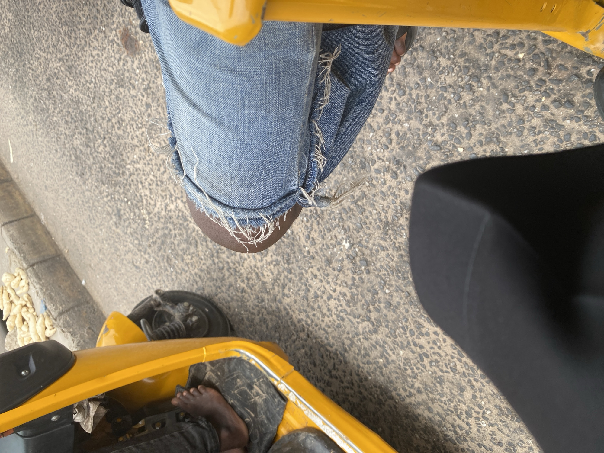 This media file shows a close-up, rotated view of a person riding on a yellow tricycle, likely in Jalingo, Nigeria. The focus is on the rider's legs, clad in distressed denim jeans, and parts of the tricycle.
The subjects are a person's legs and feet, and the yellow tricycle they are riding. The scene is set on a paved ground, possibly a street, with a concrete curb and scattered debris visible. The weather appears to be daylight, with no strong shadows indicating it's not midday.
The activity is transportation, with the person riding the tricycle. Notable details include the frayed edges and tears in the jeans, particularly around the knee area, suggesting wear and tear. The rider's bare feet are visible, one of them resting inside the tricycle. A black object, possibly a seat or a part of the tricycle's structure, is prominently visible on the right side of the frame. The tricycle itself is yellow and appears to be a common mode of transport. The background is a coarse, speckled paved surface.
There is no visible text in the image. The overall impression is a candid shot capturing a moment of everyday life and travel in an urban or semi-urban environment in Nigeria.