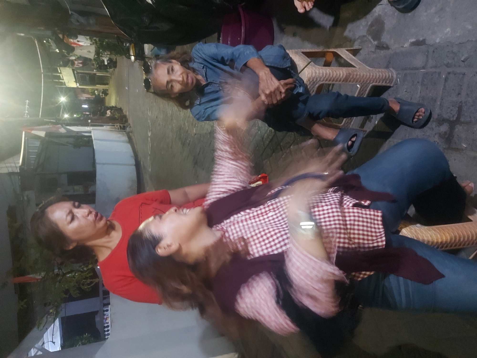 In an outdoor urban setting, possibly in Makassar, Indonesia, at night, three women are present. One woman, wearing a red t-shirt, stands and appears to be speaking. Another woman, wearing a pink and white checkered shirt and a maroon vest, is in motion, with her arm extended and her face obscured by blur. She is seated on the ground. A third woman, wearing a blue denim shirt and dark pants, sits on a small plastic stool, looking towards the viewer with her hands clasped. The environment is a narrow alleyway with buildings on either side. There are lights strung across the alley and clothes hanging to dry. A large purple tub is visible behind the woman on the stool. The ground is paved with cobblestones.