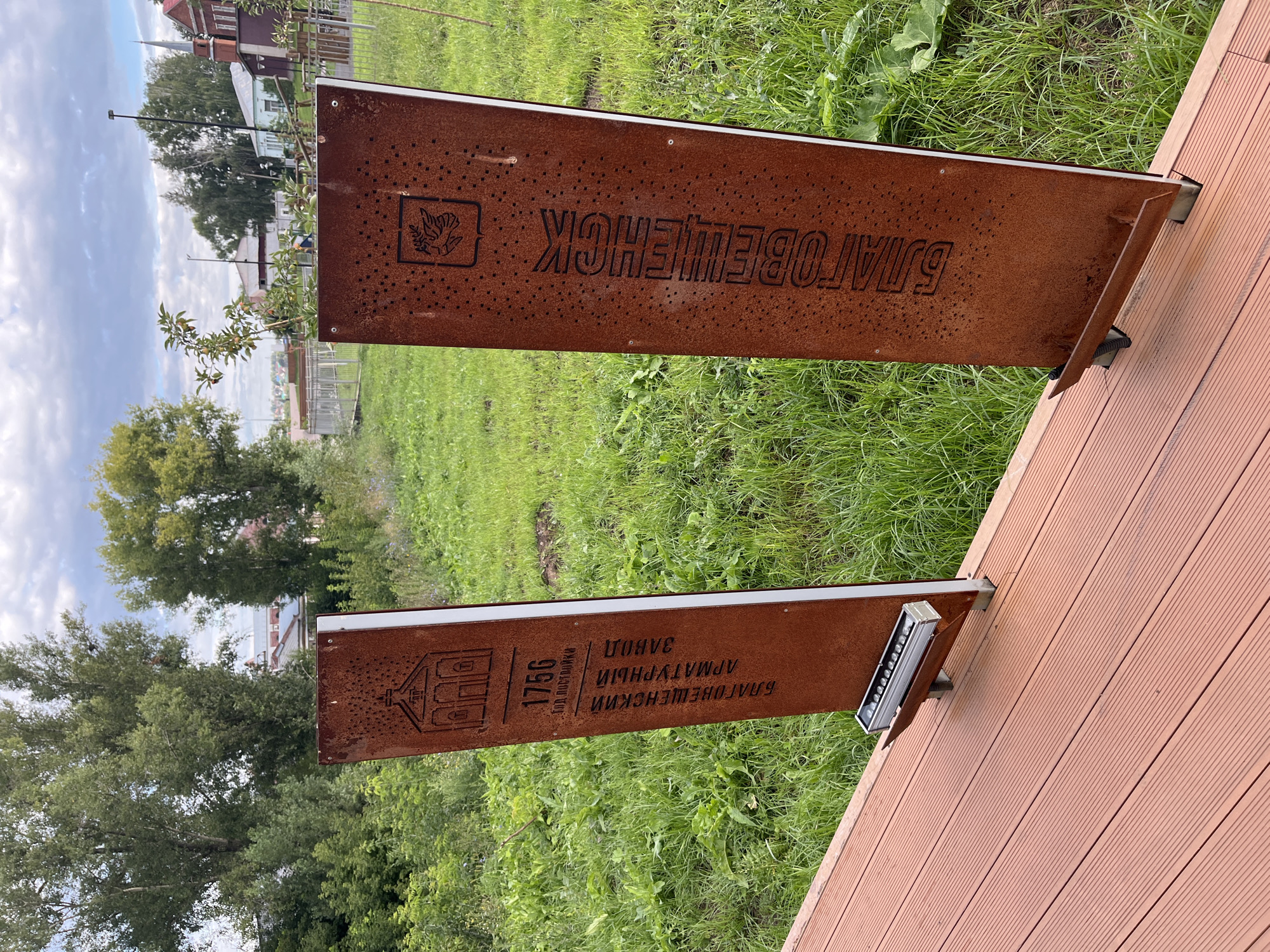 Two weathered, rusted metal panels stand upright on a wooden deck, set against a backdrop of green grass and trees under a cloudy sky. The panel on the left features a graphic of a building and text in Russian that reads "1756 ГОД ПОСТРОЙКИ БЛАГОВЕЩЕНСКИЙ АРМАТУРНЫЙ ЗАВОД" (1756 YEAR OF CONSTRUCTION BLAGOVESHCHENSK ARMATURE FACTORY). Below this text, a row of small lights is affixed to the panel. The panel on the right displays a coat of arms and the word "БЛАГОВЕЩЕНСК" (BLAGOVESHCHENSK) in large, Cyrillic lettering. The environment suggests an outdoor park or public space within the city of Blagoveshchensk, Russia, likely commemorating historical industrial sites. There are no people or animals visible in the frame.