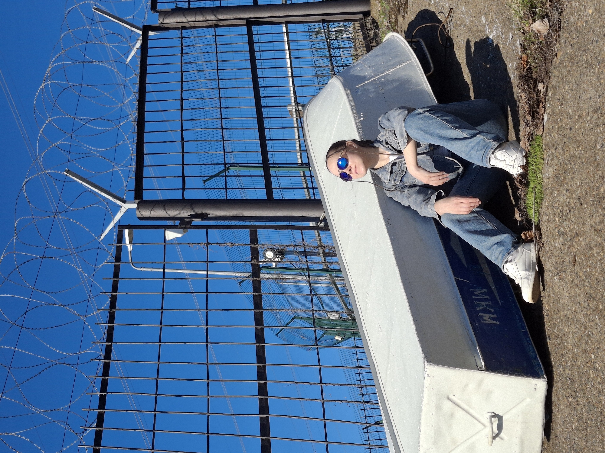 The image depicts an outdoor scene in Chaikovsky, Russia, under a clear, bright blue sky. A young person is seated casually on what appears to be an upturned, light-colored aluminum boat. They are dressed in a denim jacket, blue jeans, and white sneakers, wearing reflective blue mirrored sunglasses. Their hands are clasped loosely in their lap, and they have braided hair visible.
The boat has the letters "MKM" stenciled on its side in a dark blue or black color. In the background, a tall metal grid fence is prominently featured, topped with multiple coils of razor wire, also known as concertina wire, indicating a secure or restricted area. The ground beneath the boat is concrete, with some dry foliage visible along its edge.
The bright sunlight creates long, distinct shadows on the ground, suggesting it is either mid-morning or late afternoon on a sunny day. The overall atmosphere is one of relaxed observation, contrasting the casual posture of the person with the stark, utilitarian elements of the fence and barbed wire.