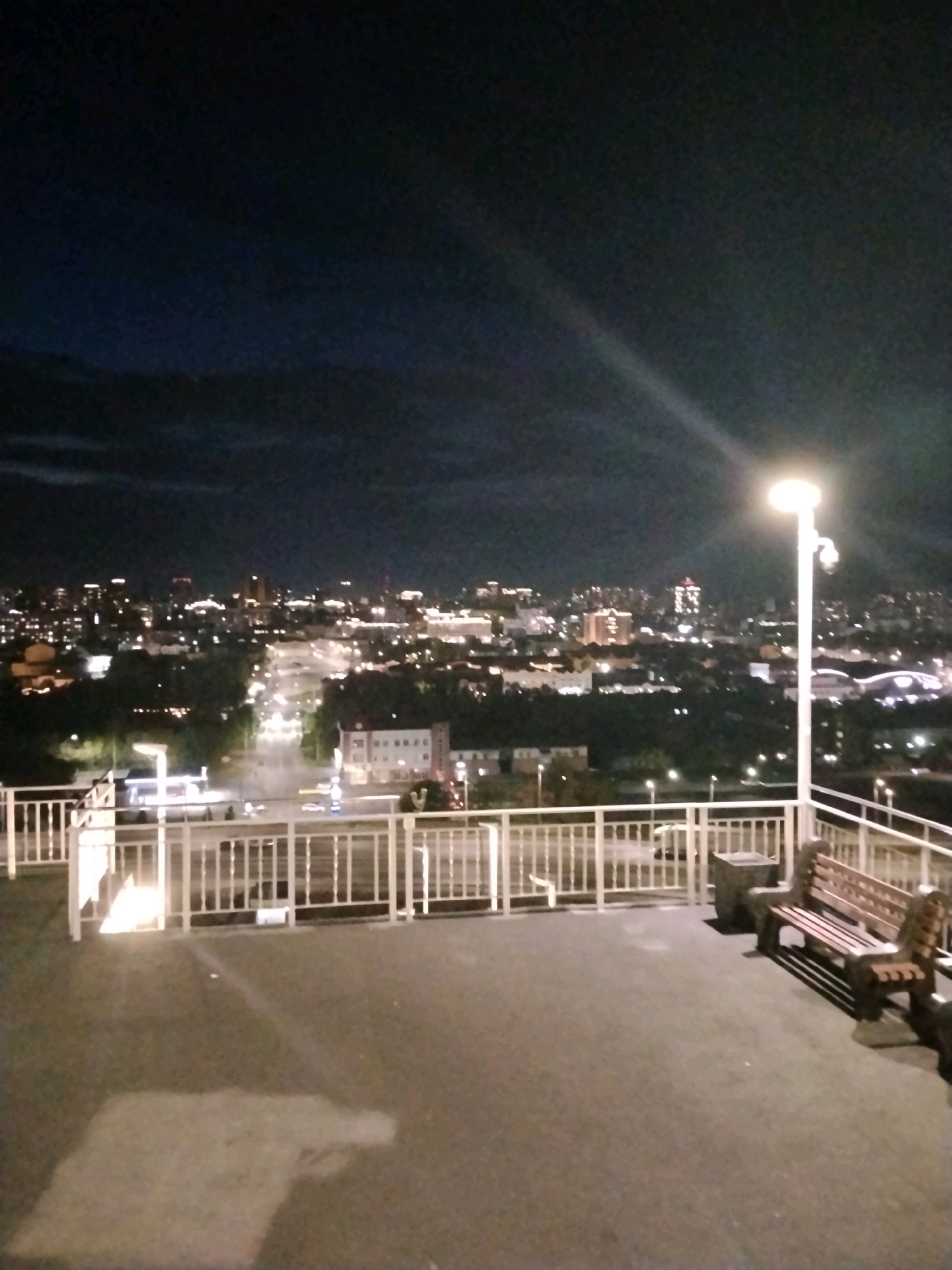 The image depicts a nighttime view of a city, identified as Barnaul, Russia. In the foreground, there is a paved area with a white railing. A wooden bench and a trash receptacle are situated on this paved area, next to the railing. A tall street lamp with a bright light stands to the right, illuminating the immediate surroundings. Beyond the railing, a sprawling cityscape is visible, characterized by numerous lights from buildings and vehicles on roads. The sky above is dark with some clouds. The scene appears to be an overlook or a balcony offering a panoramic view of the city at night.