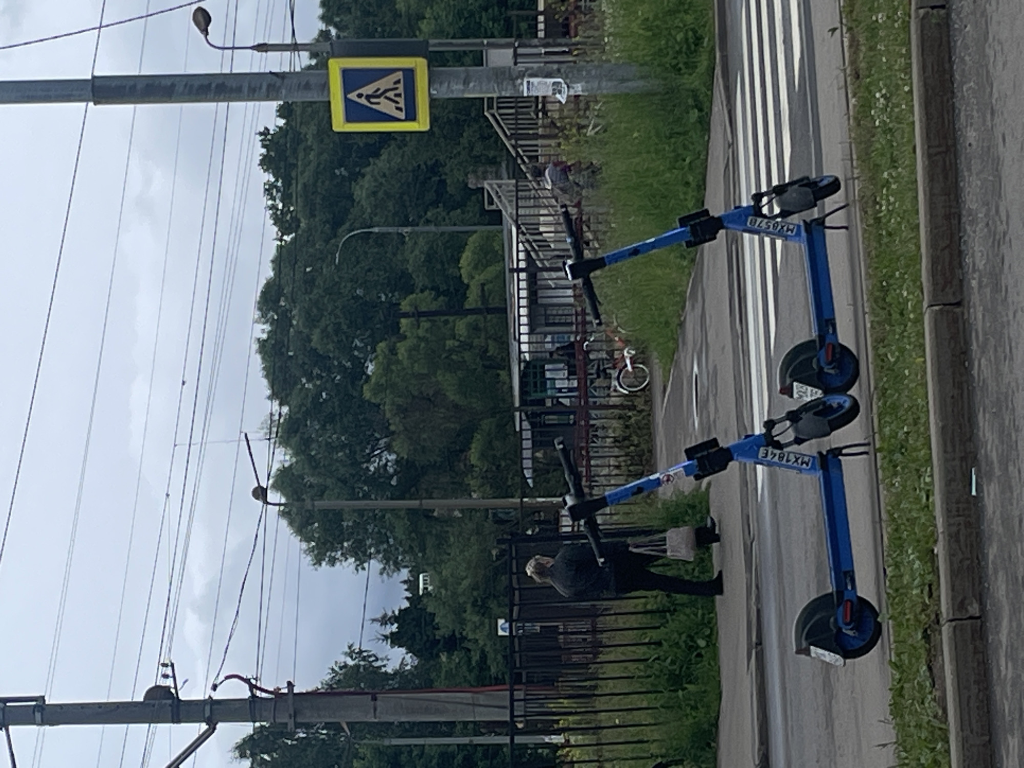The image shows an outdoor scene in Рассудово, Russia. Two electric scooters, one blue and one dark, are parked on the side of a road next to a crosswalk. The blue scooter has the text "MXG578" on it, and the other scooter has "MX184E". In the background, there is a pedestrian crossing sign, a metal fence, and lush green trees. A person in dark clothing is partially visible, appearing to be walking away from the scooters. Overhead power lines are prominent in the sky, which is overcast, suggesting a cloudy day. The road surface is visible, as is a strip of grass with small white flowers bordering the road. The scene appears to be a public area, possibly a park or a street with pedestrian amenities.