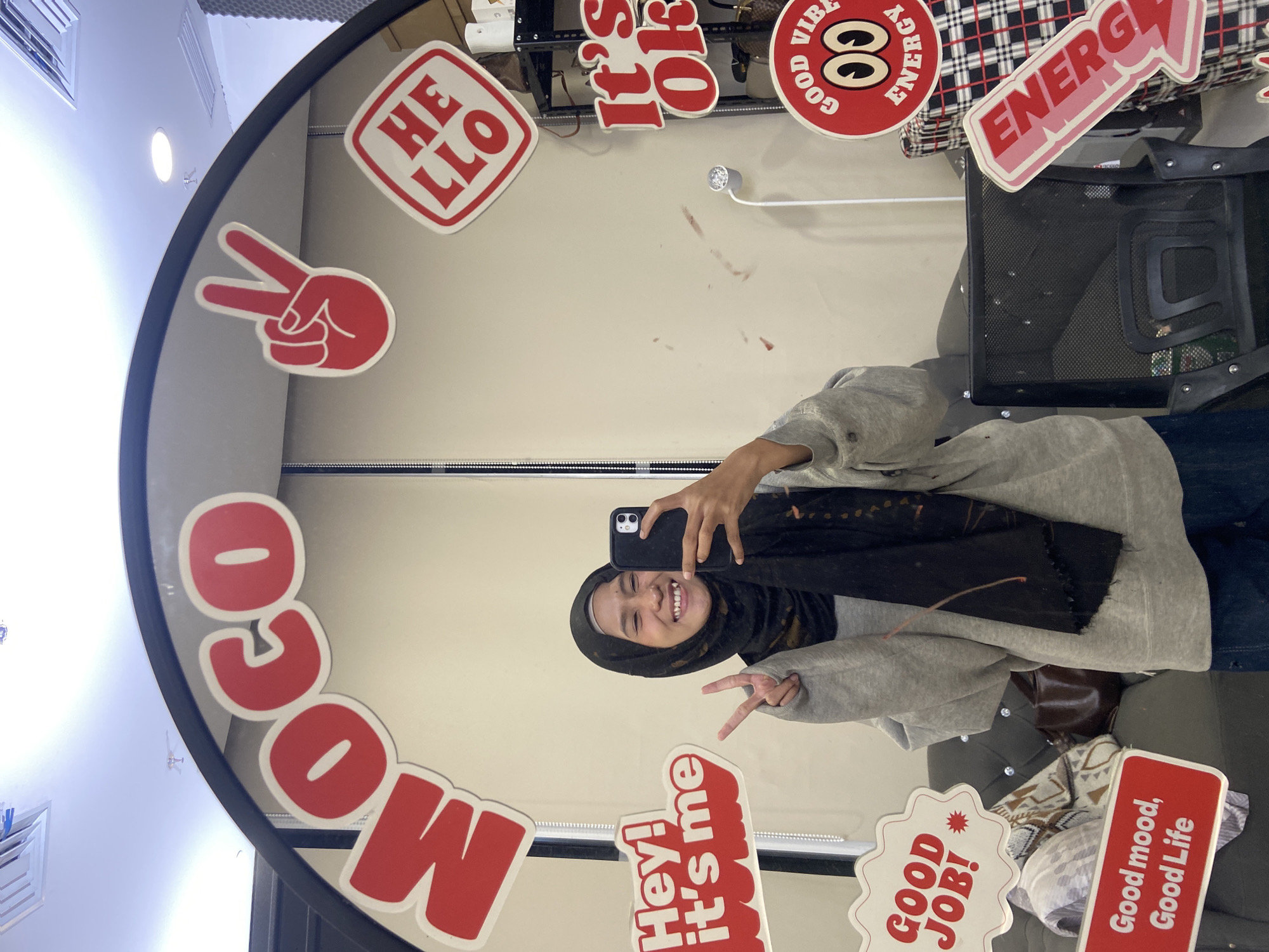 A young woman wearing a hijab stands in front of a mirror taking a selfie in North Jakarta, Indonesia. Various red and white signs with encouraging phrases such as "Hello," "It's Ok," "Good Vibes," "Energy," "Moco," "Hey! It's me," "Good Job!" and "Good mood, Good Life" surround the mirror's edge. The woman is smiling and making a peace sign. Behind her, there is a gray chair, a checkered cloth, and a black metal rack.