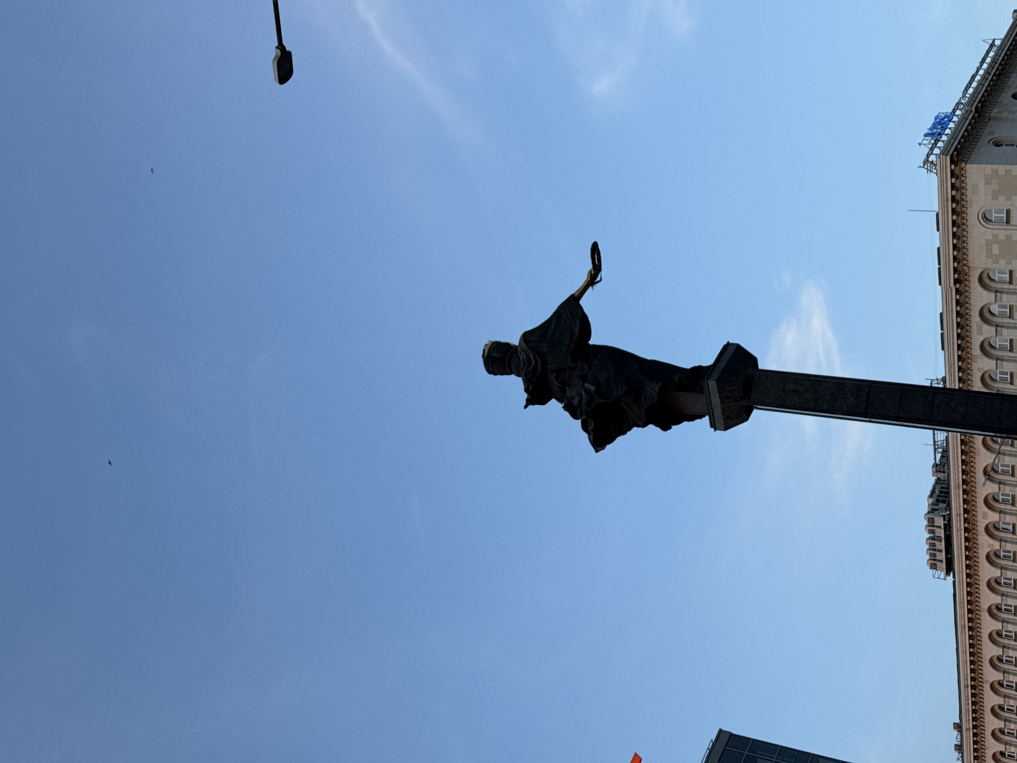 The image, when viewed upright, displays the Monument to Saint Sofia on a tall column, set against a clear blue sky in Sofia, Bulgaria. The statue, made of dark material (likely bronze), depicts a draped female figure holding a wreath in her raised right hand and an owl, symbolizing wisdom, on her left arm.
To the right of the statue, a large, light-colored building with intricate architectural details is visible. It features numerous arched windows and decorative elements along its facade. On the top of this building, a blue sign clearly spells out "SHERATON," identifying it as the former Sheraton Sofia Hotel Balkan (now Hotel Balkan), which stands adjacent to the monument.
The sky is a vibrant, cloudless blue, indicating clear and sunny weather. A dark street light fixture is present in the upper left of the frame. A small, dark bird can be seen flying in the distant sky to the left. The time of day appears to be bright daylight, likely morning or afternoon, given the even illumination. The scene captures an iconic landmark and its urban surroundings, with no specific activity taking place beyond the static display of the monument and building.