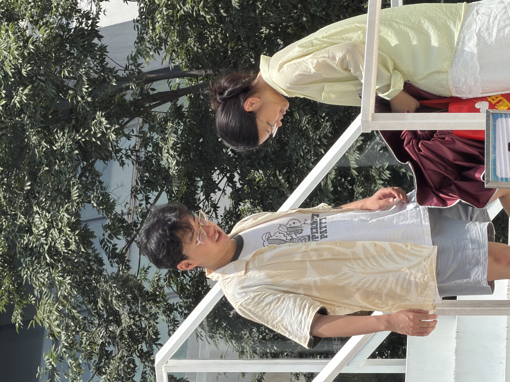 Two young adults, a man and a woman, are standing on a white metal staircase outdoors. The man, on the left, wears a light-colored, short-sleeved button-down shirt with a tropical leaf pattern over a white t-shirt and light gray shorts. He has dark hair, wears glasses, and is looking down and to his right. The woman, on the right, wears a light green, long-sleeved translucent shirt over a white skirt with lace trim. Her dark hair is pulled back in a ponytail, and she wears glasses. She is looking down at a dark red umbrella and a red tote bag. The background consists of lush green foliage from a tree and a building. The overall impression is a casual outdoor setting, possibly in a park or urban area with greenery.