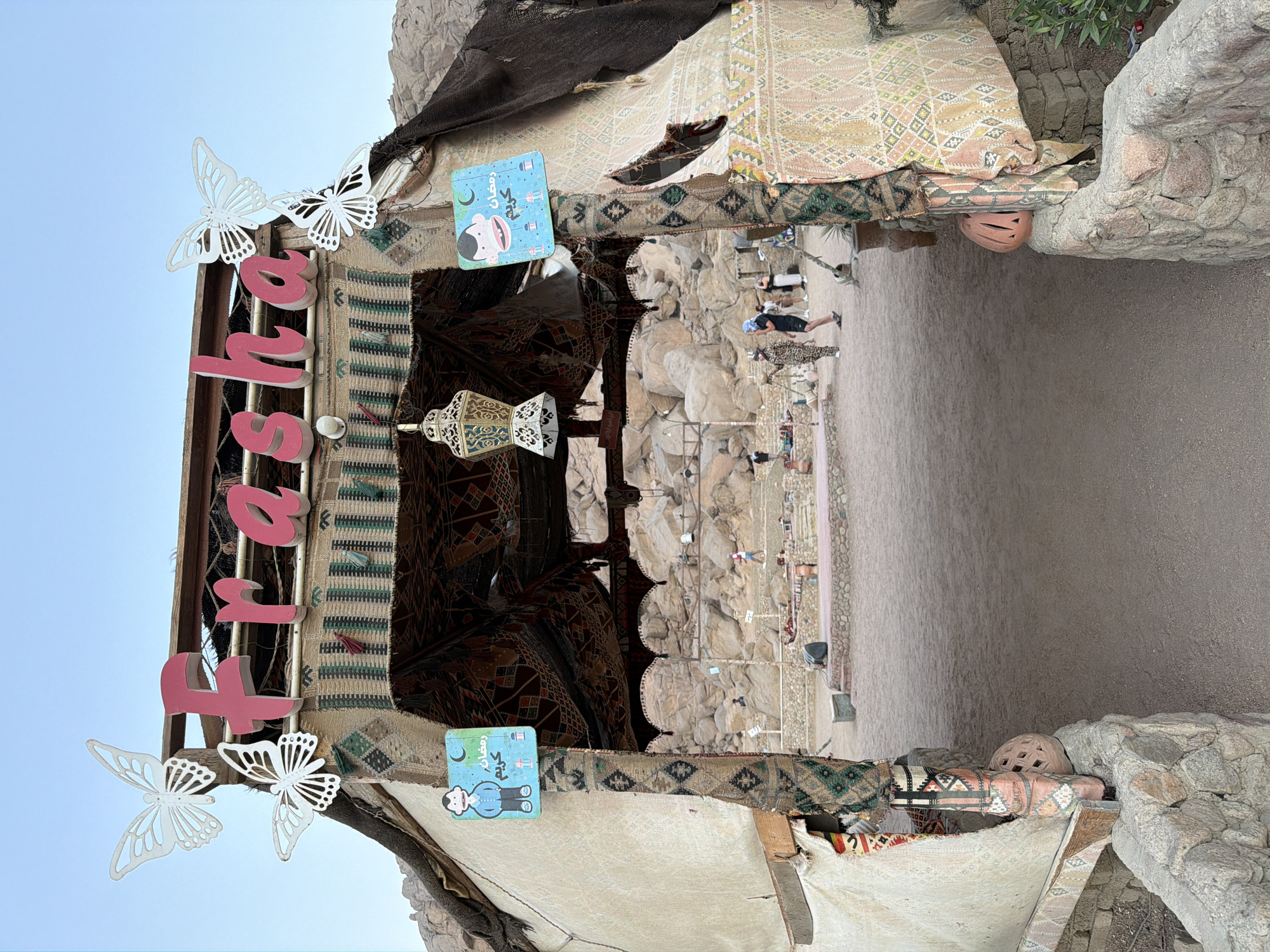 The image shows an outdoor seating area within a desert landscape, likely in Sharm El Sheikh, Egypt, as indicated by the Ramadhan decorations. The entrance to the seating area is adorned with pink lettering spelling "Frasha" and two white butterfly cutouts. Colorful textiles hang from the roof structure, and ornate lanterns are suspended above.
In the background, several people are visible, some walking and others seated. The ground is sandy, and large rocks are scattered throughout the area, typical of a desert environment. The overall scene suggests a relaxed, open-air dining or gathering space decorated for a special occasion.